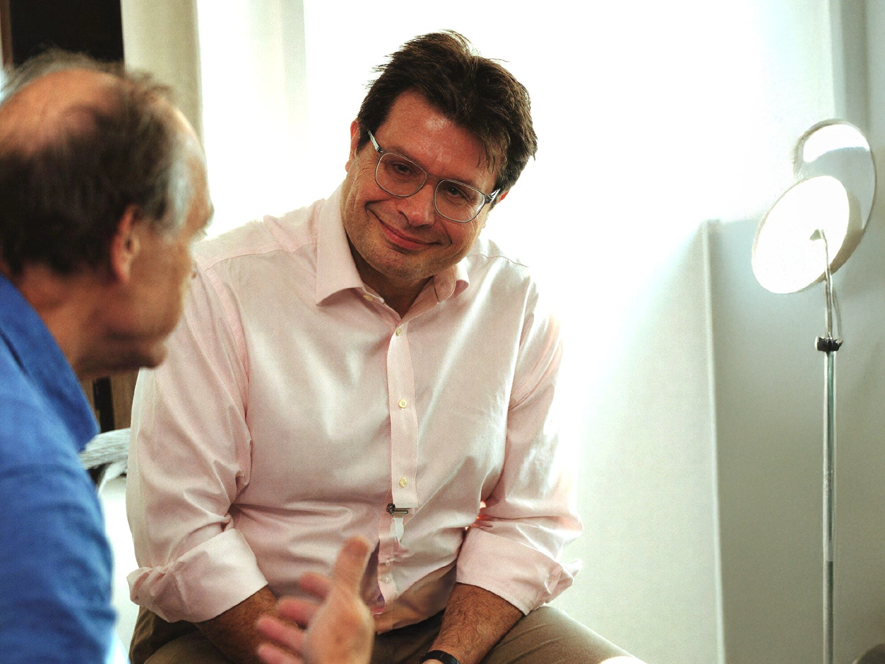Man in glasses and pink shirt attentively listening to another man in a blue shirt during a conversation.