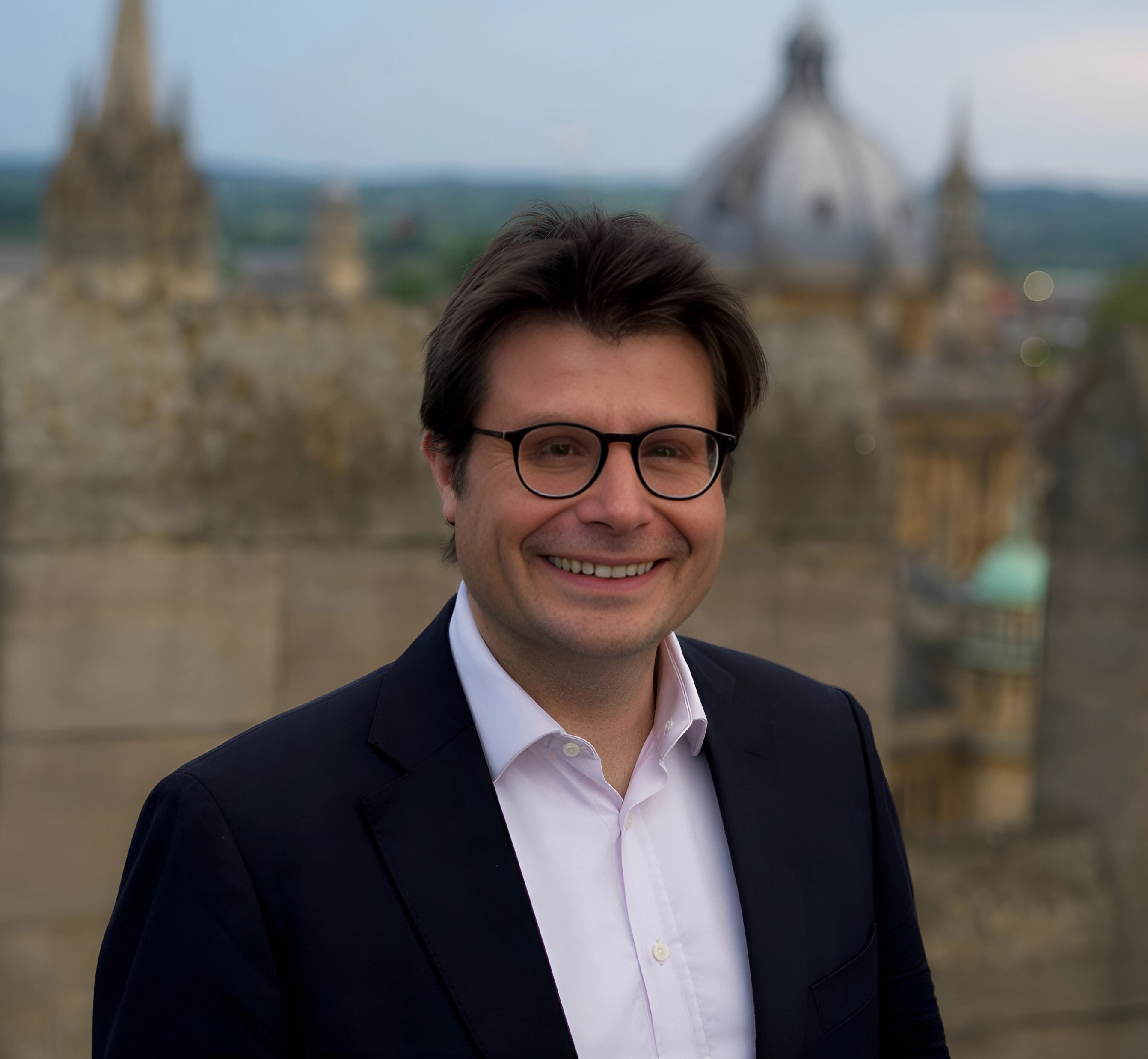 Smiling man with glasses in a dark suit and white shirt outdoors with a blurred cityscape background.