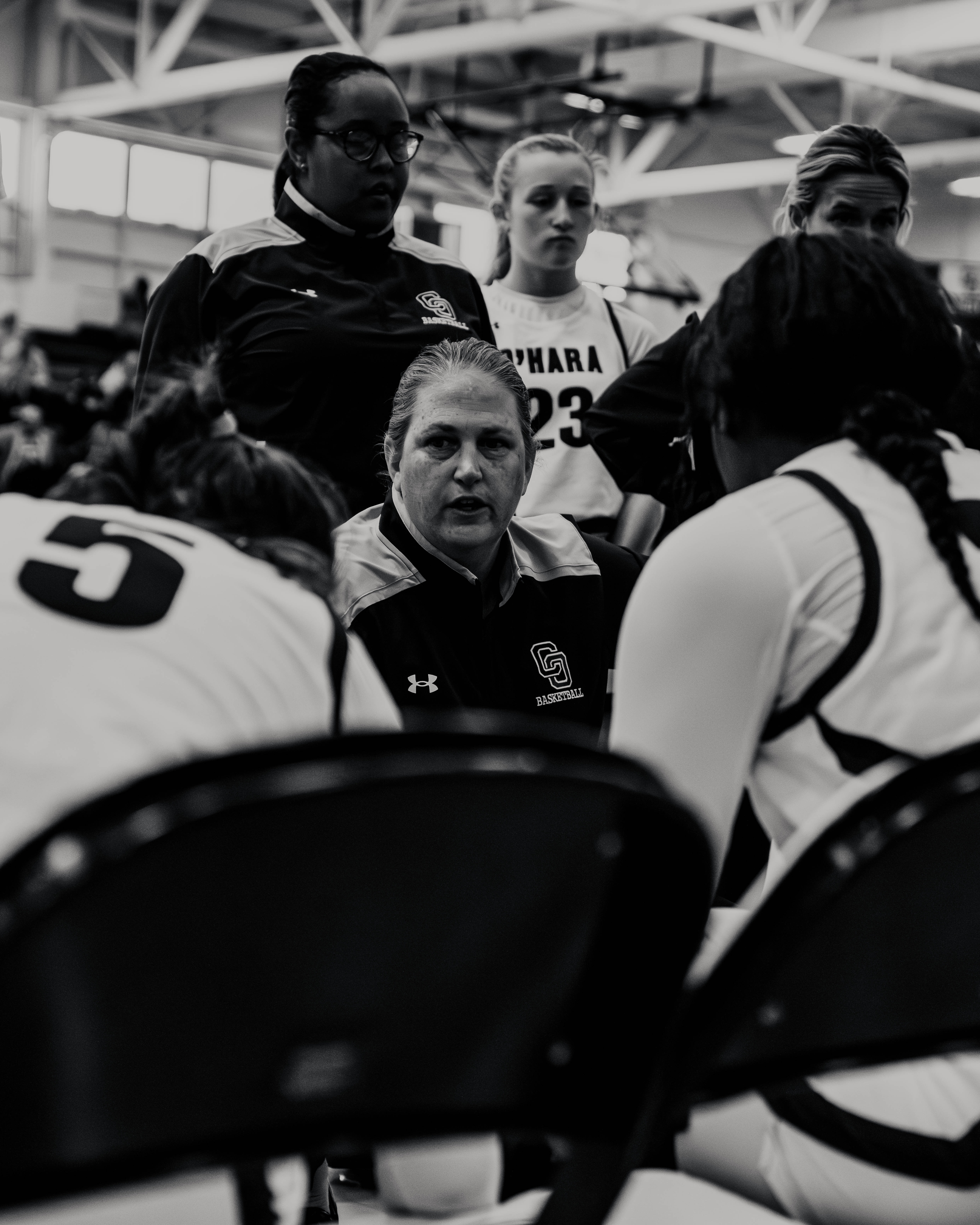 Coach giving instructions to female basketball players during a game timeout in a gym.
