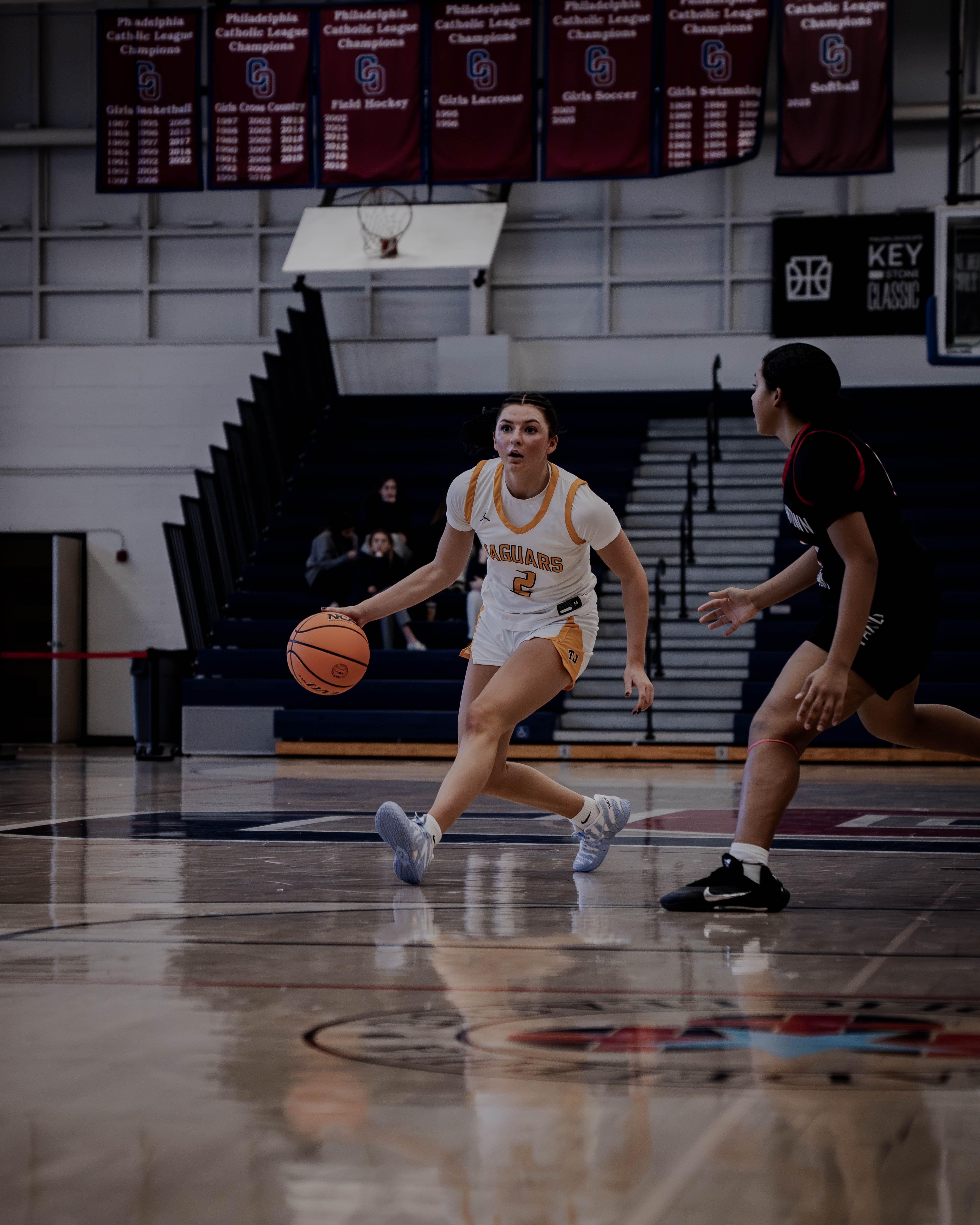 Female basketball player in white and yellow Jaguars uniform dribbling the ball while being defended by another player in black uniform.