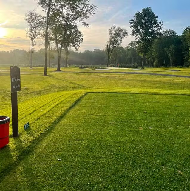 Early sunlit golf course tee area with a sign indicating Hole 6 at 109 yards, surrounded by trees and a clear sky.