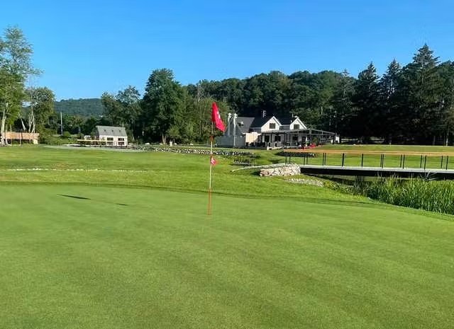 Golf green with a red flag on a sunny day, clubhouse and trees in the background.