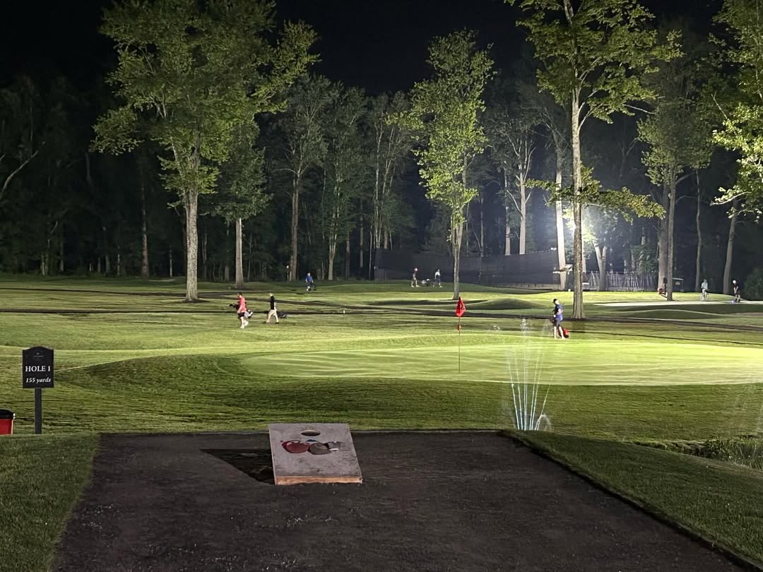 Nighttime golf course scene with a putting green, red flag, sprinklers on, and people walking and playing in the background.