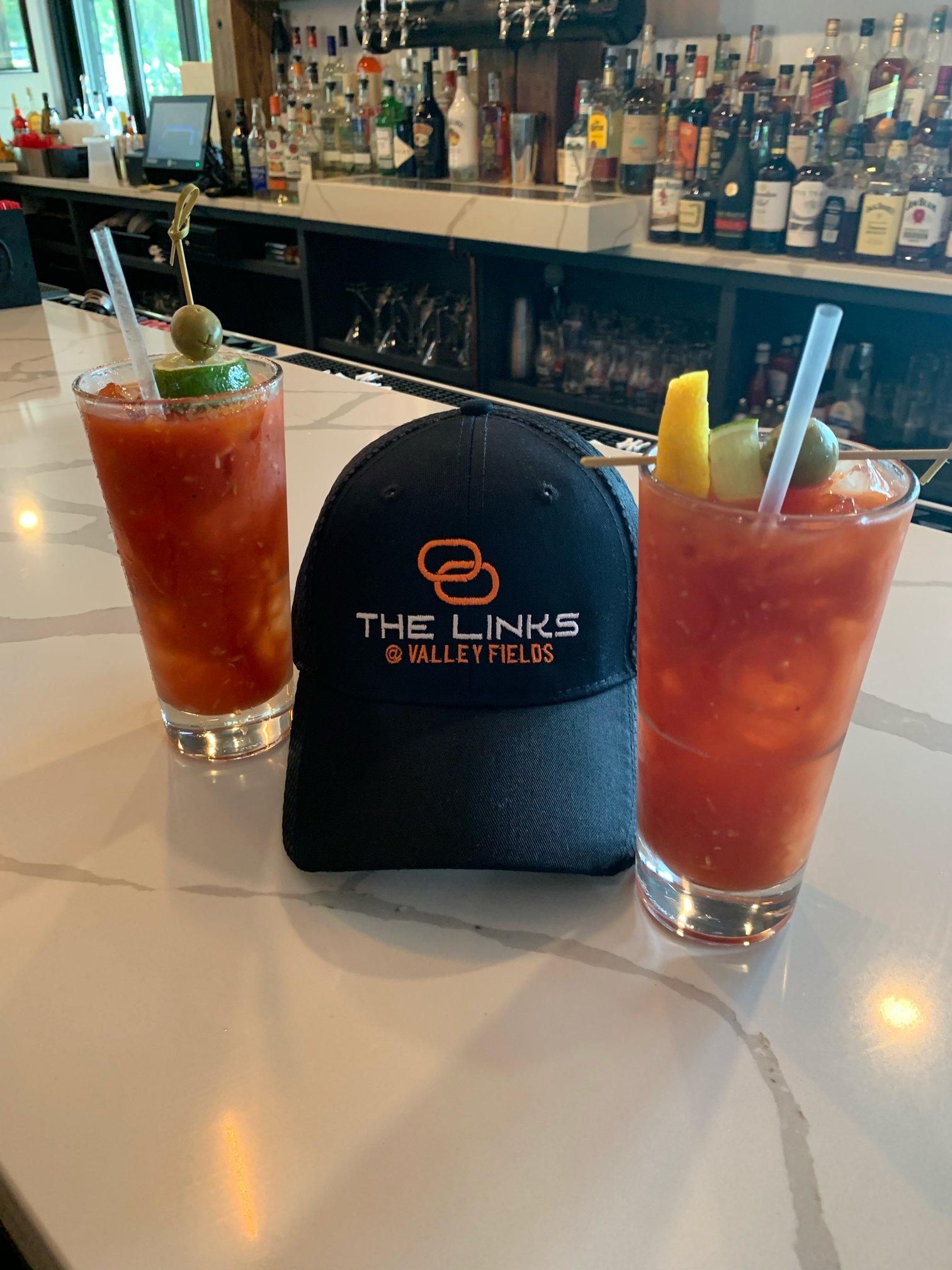 Two Bloody Mary cocktails garnished with olives and lime or lemon wedges flanking a black cap embroidered with 'The Links @ Valley Fields' on a white marble bar counter with liquor bottles in the background.