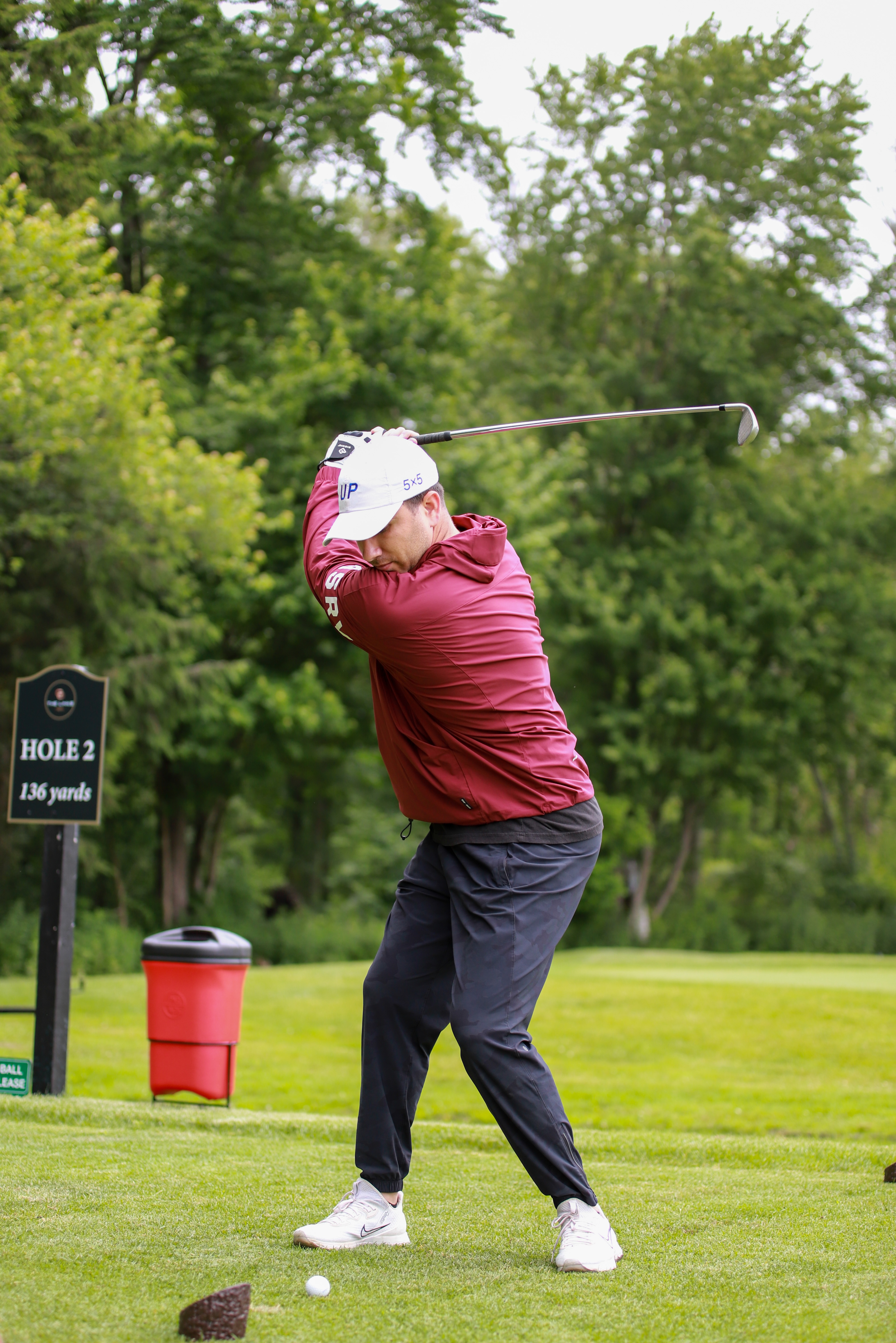 Golfer in a red jacket and white cap swinging a golf club on the tee of hole 2 with a sign showing 136 yards.