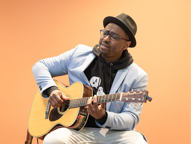 Parfait Bassalé singing with his guitar on an orange background