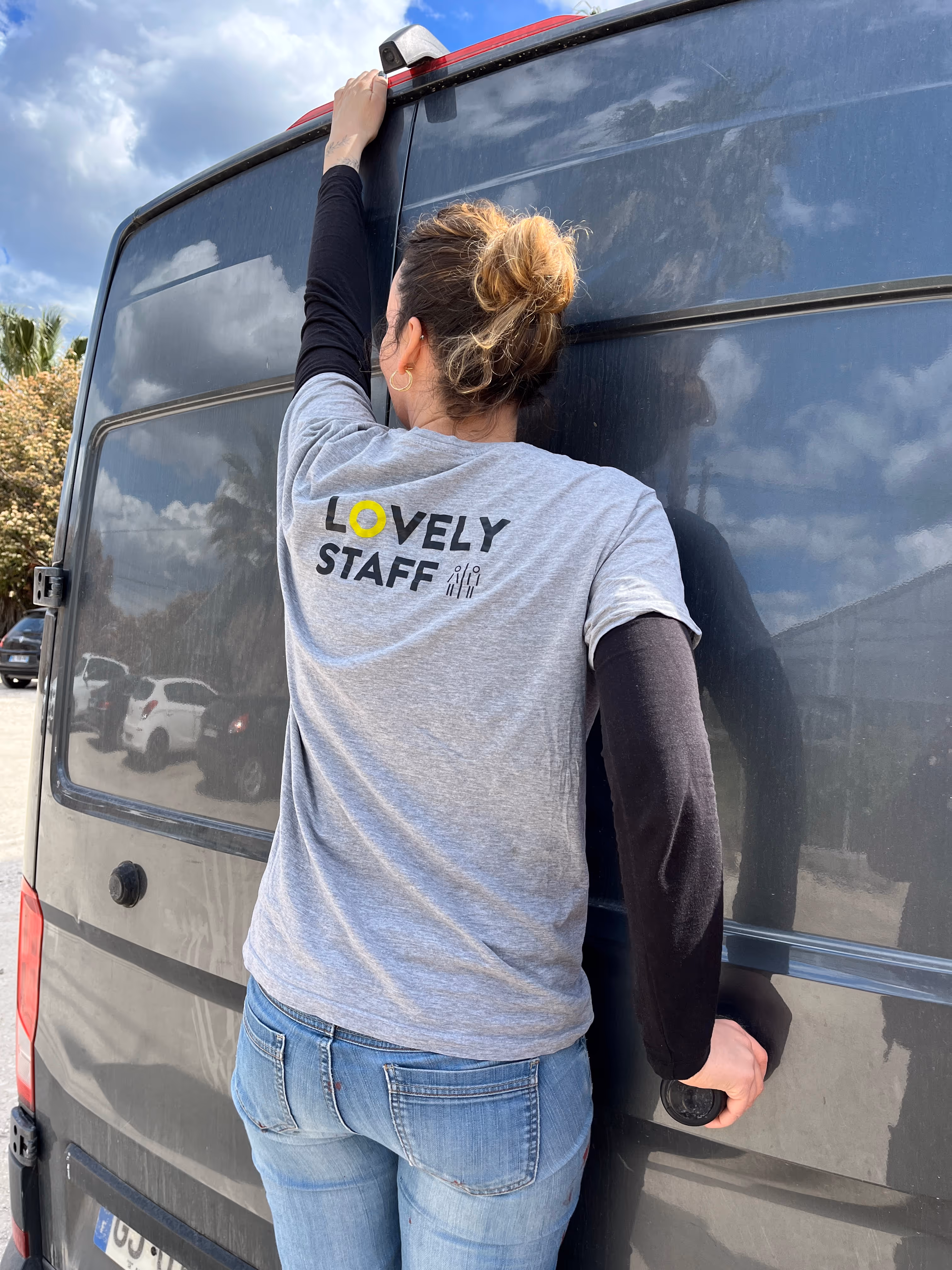 Femme vue de dos portant un t-shirt gris avec l'inscription 'LOVELY STAFF' et un long sous-pull noir, tenant la porte arrière d'une grande camionnette noire.