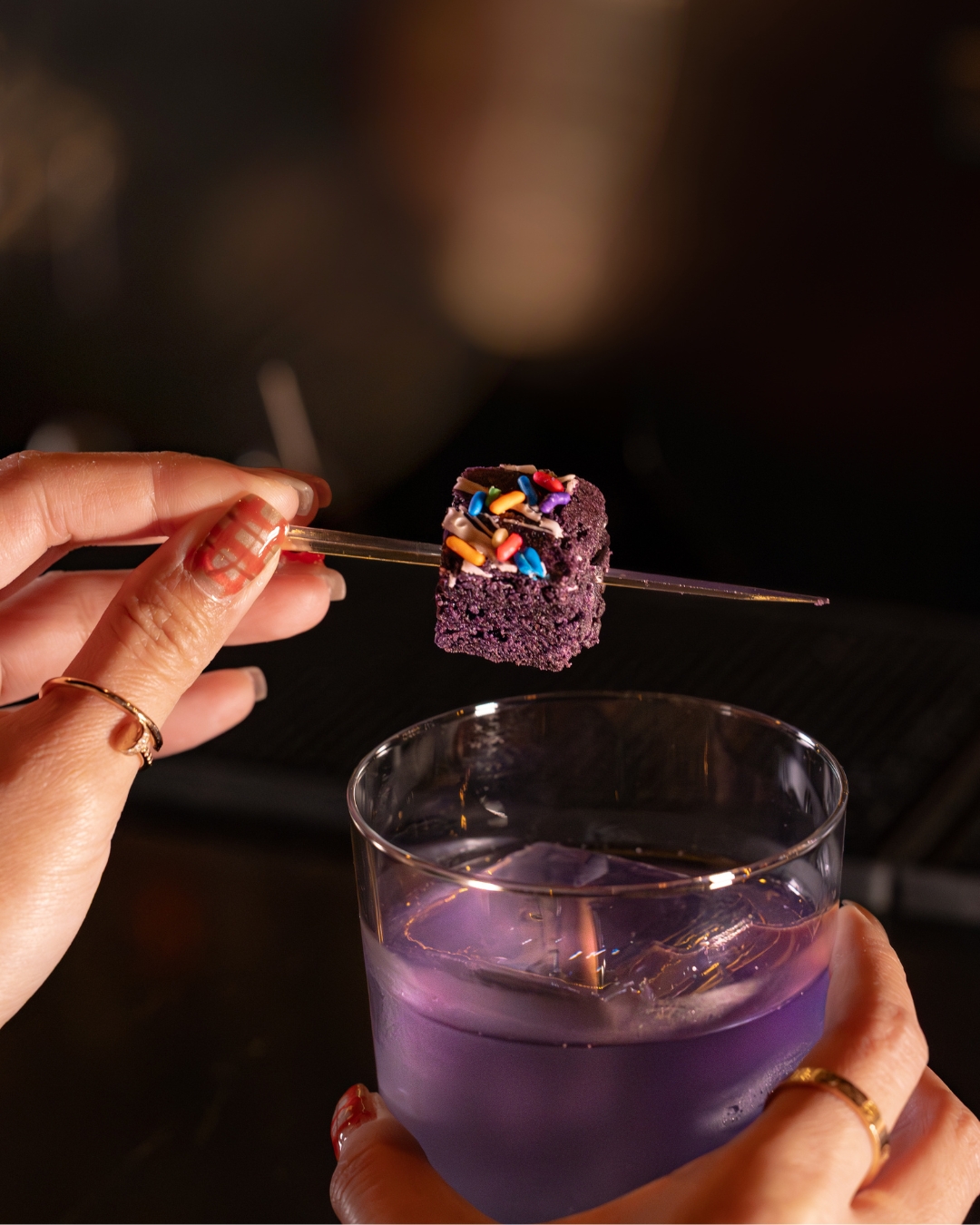 Hand holding a cocktail glass filled with a purple drink and a stick with a purple cake cube topped with colorful sprinkles.