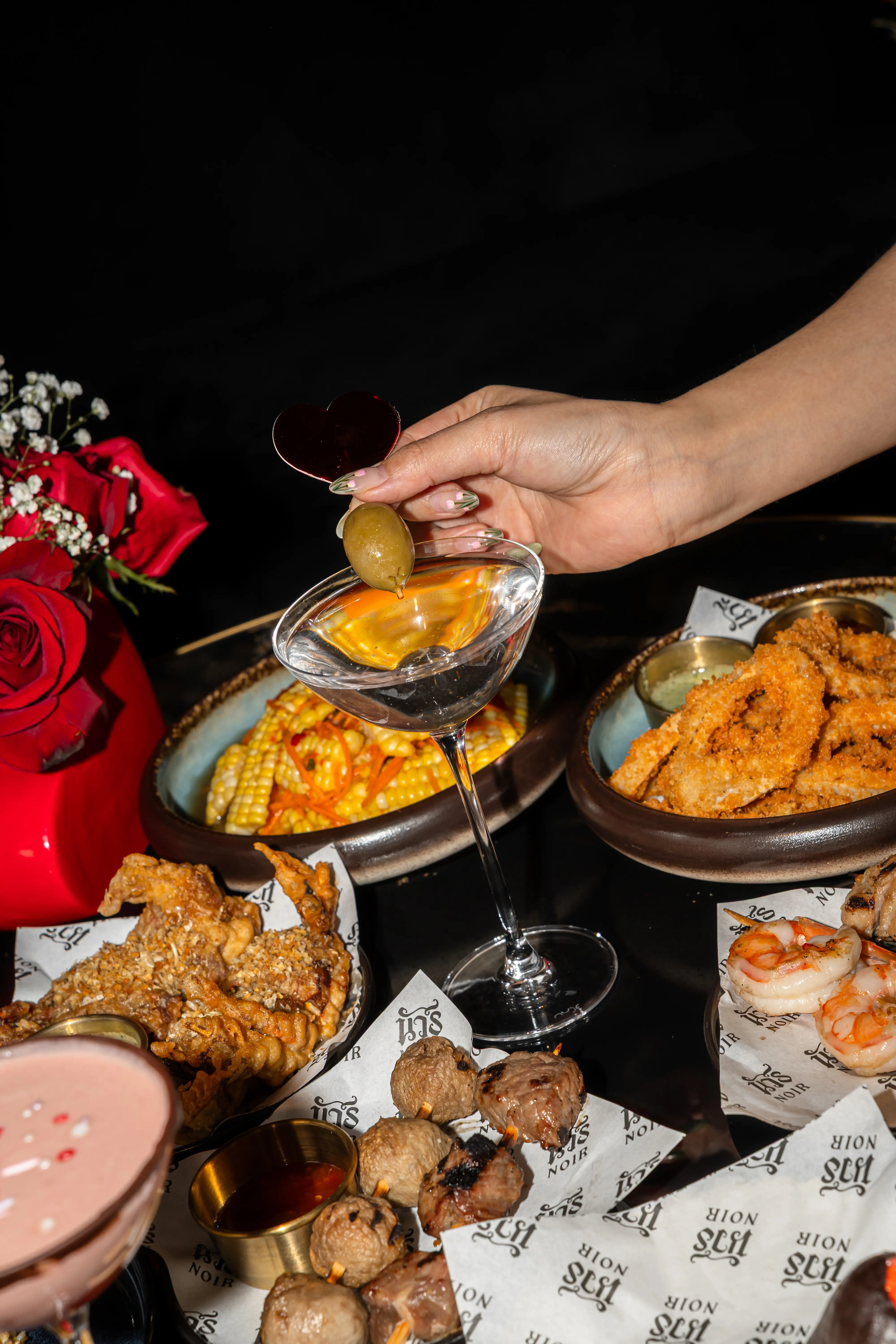 Hand holding a martini glass with an olive garnish above a table filled with various dishes including fried onion rings, grilled shrimp, skewered meat, and corn salad, with a red vase of roses in the background.