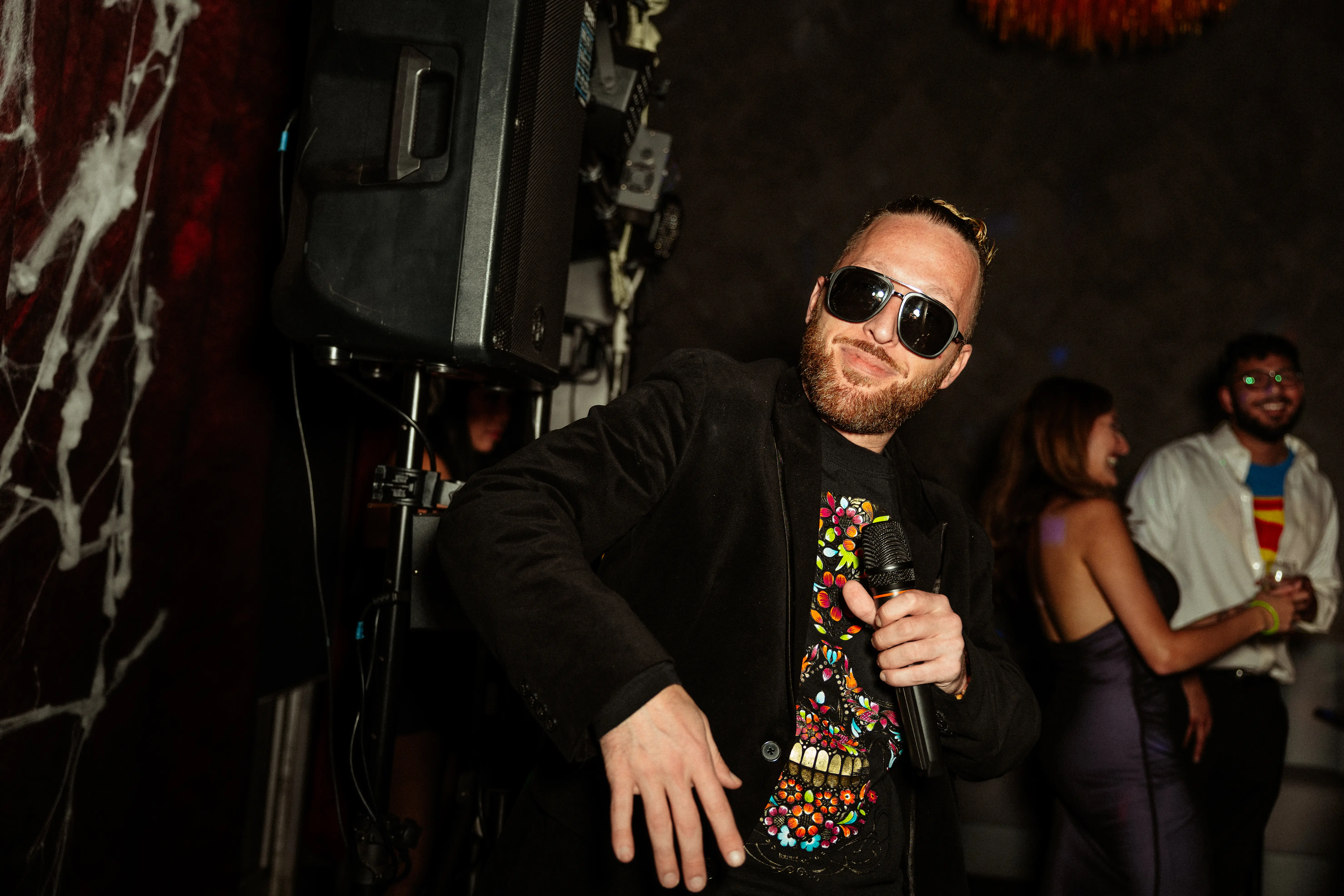 Man wearing sunglasses and a black jacket with a colorful skull shirt holding a microphone at a party.
