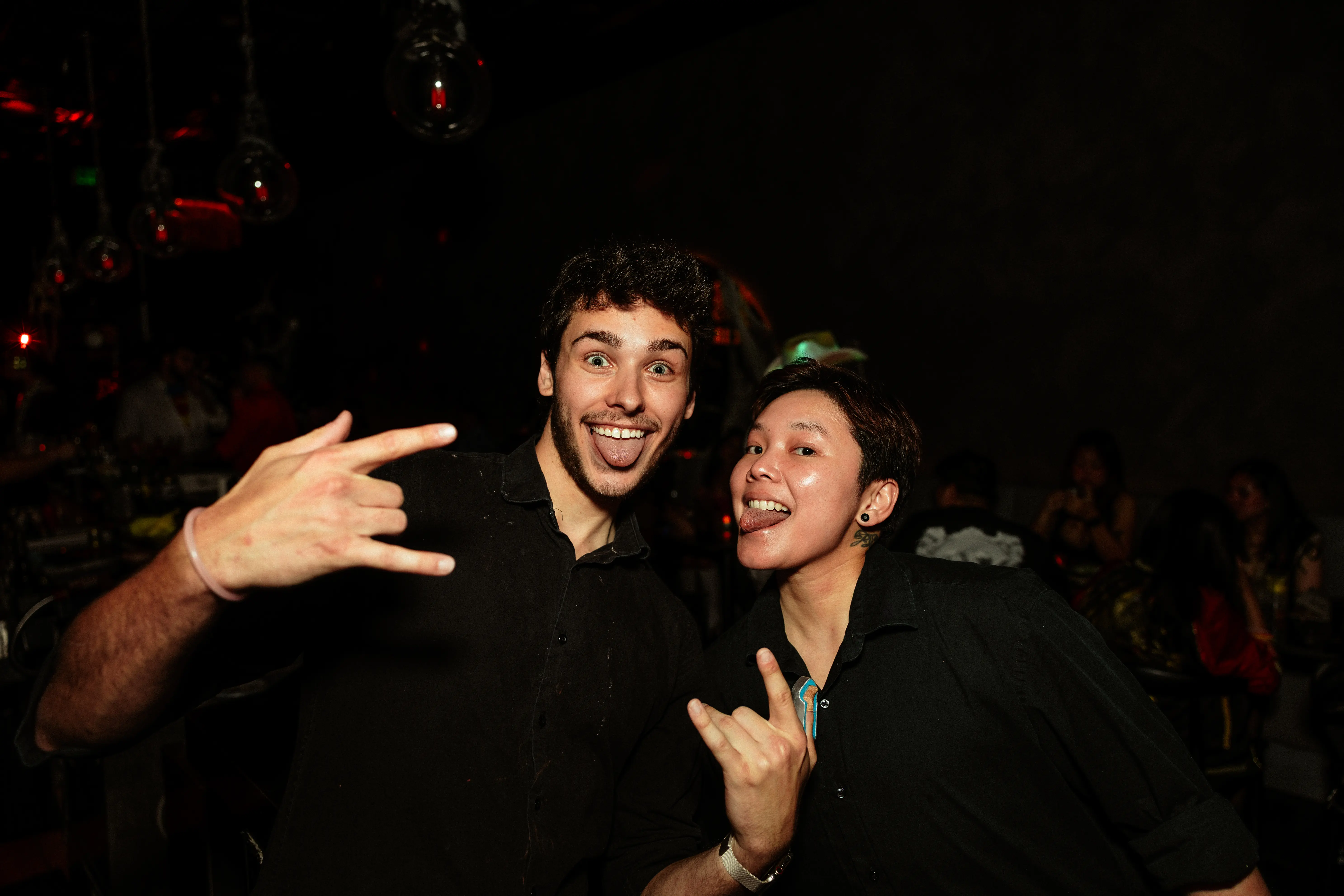 Two young men in black shirts making rock hand signs and sticking out their tongues in a dimly lit party setting.