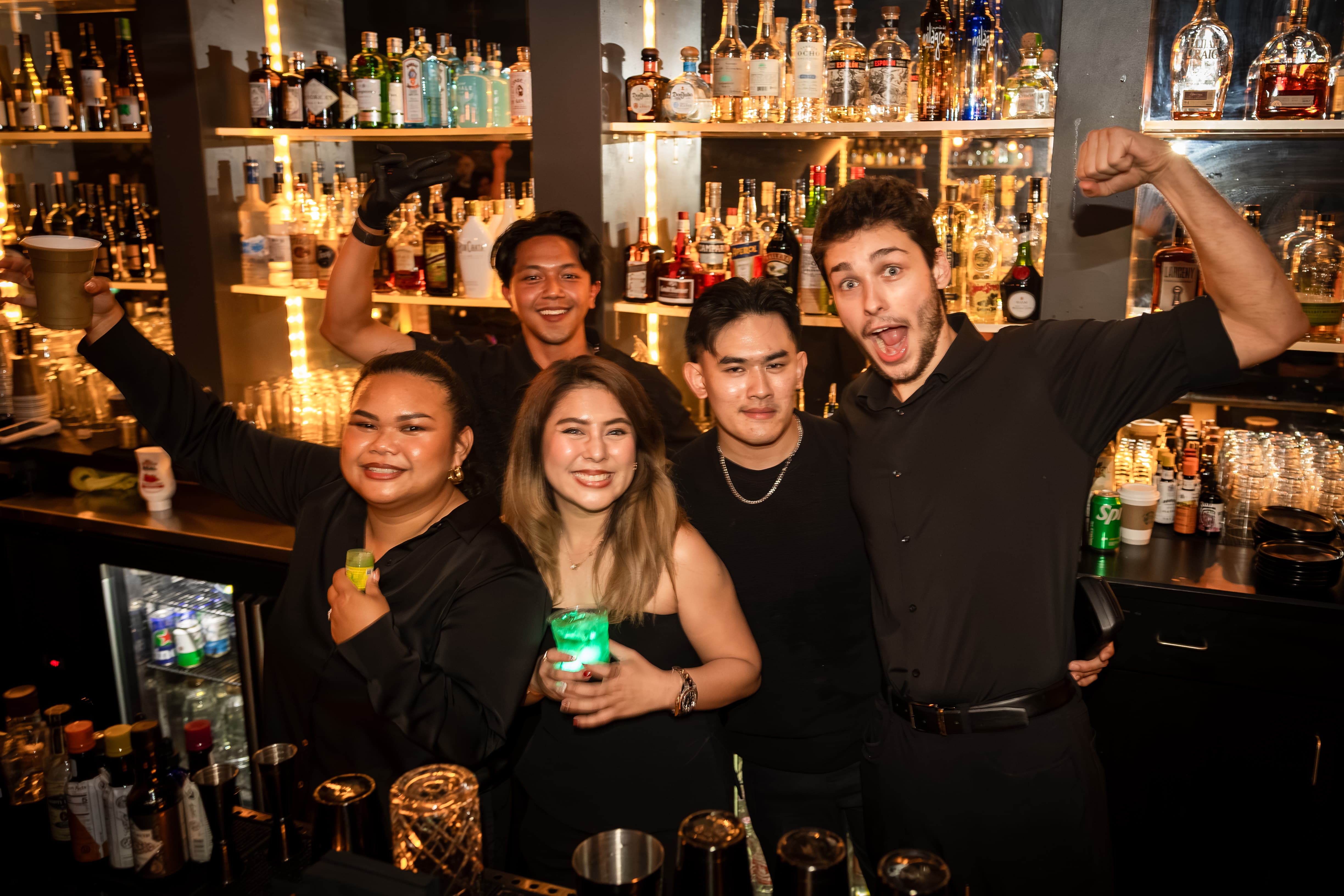 Five bar staff smiling and posing behind a bar counter with bottles and glasses on shelves in the background.