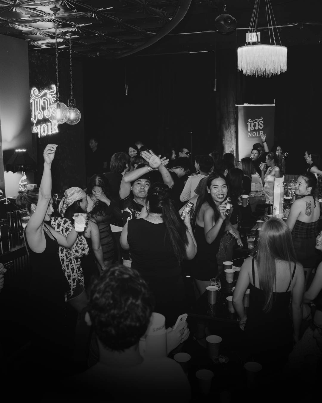 Crowd of people enjoying a lively night at a bar with drinks and socializing under decorative lighting.