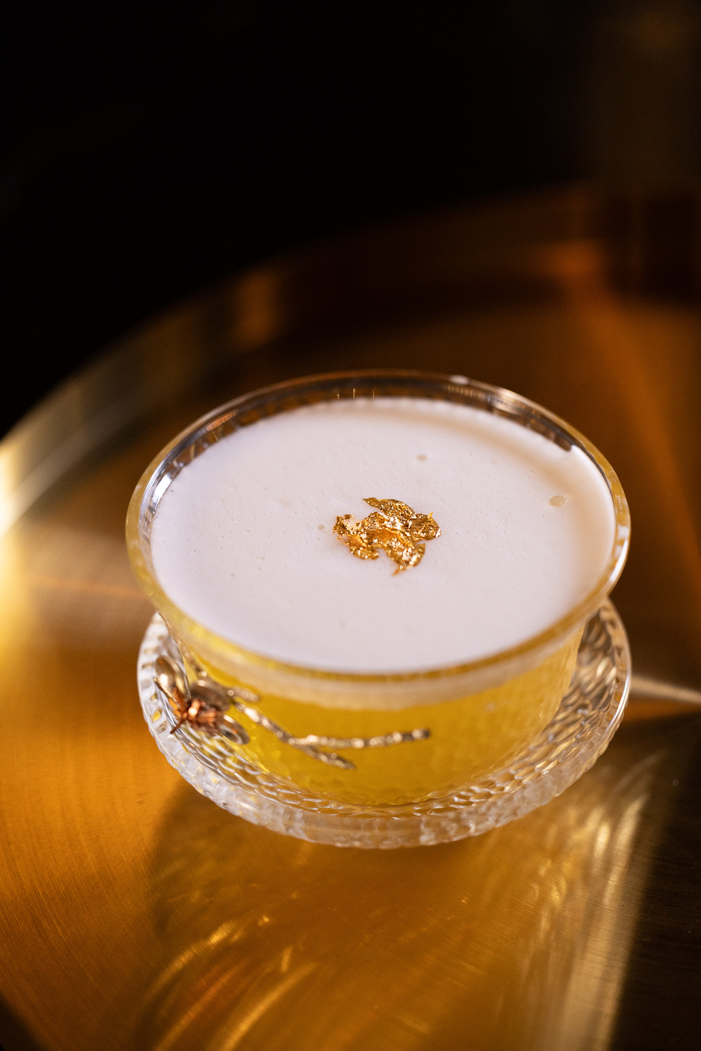 Yellow cocktail with white foam and gold leaf garnish in an ornate glass cup on a metallic gold surface.
