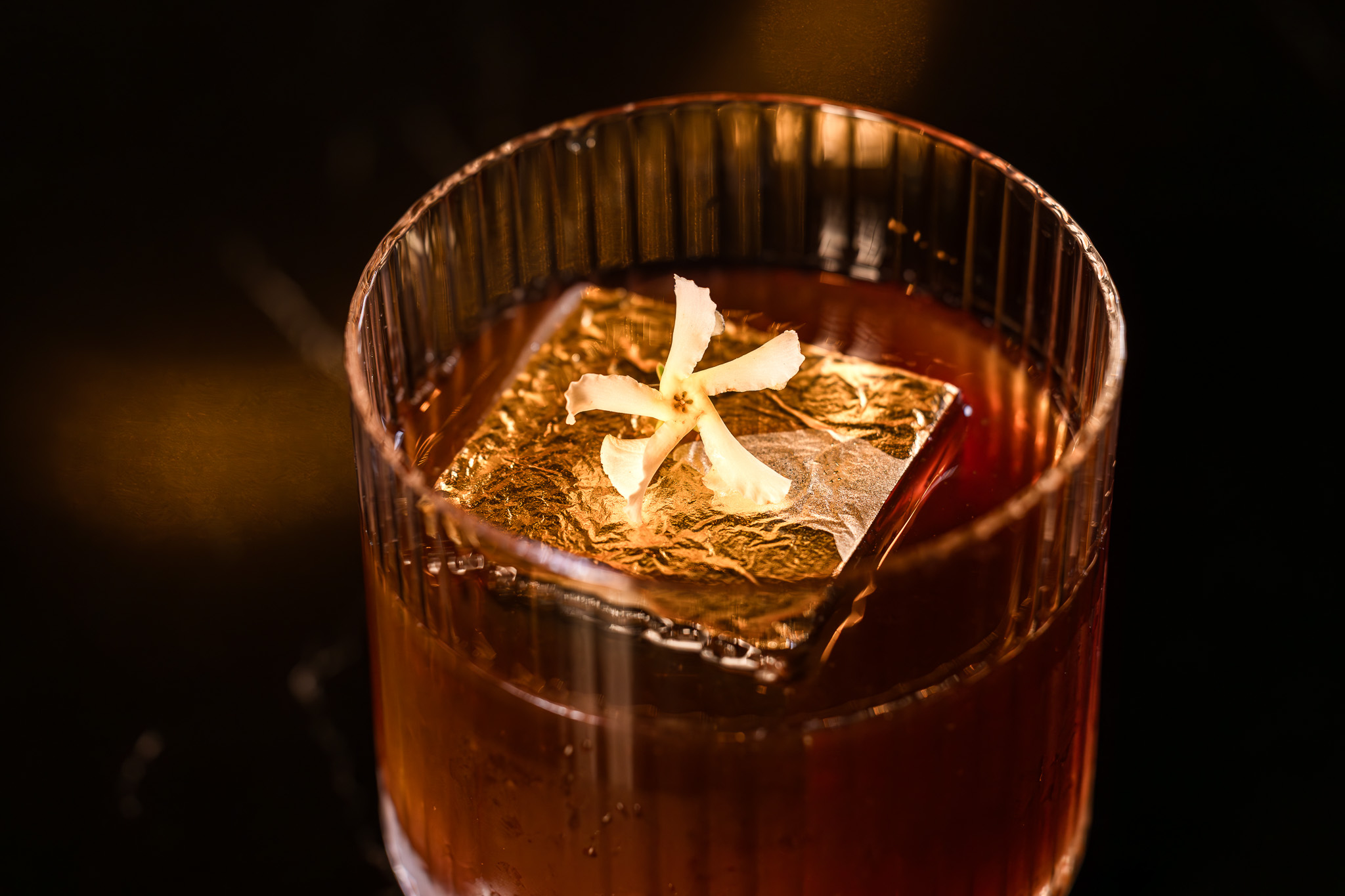 Close-up of a dark amber cocktail in a ribbed glass with a large gold-foil-covered ice cube and a small white flower on top.