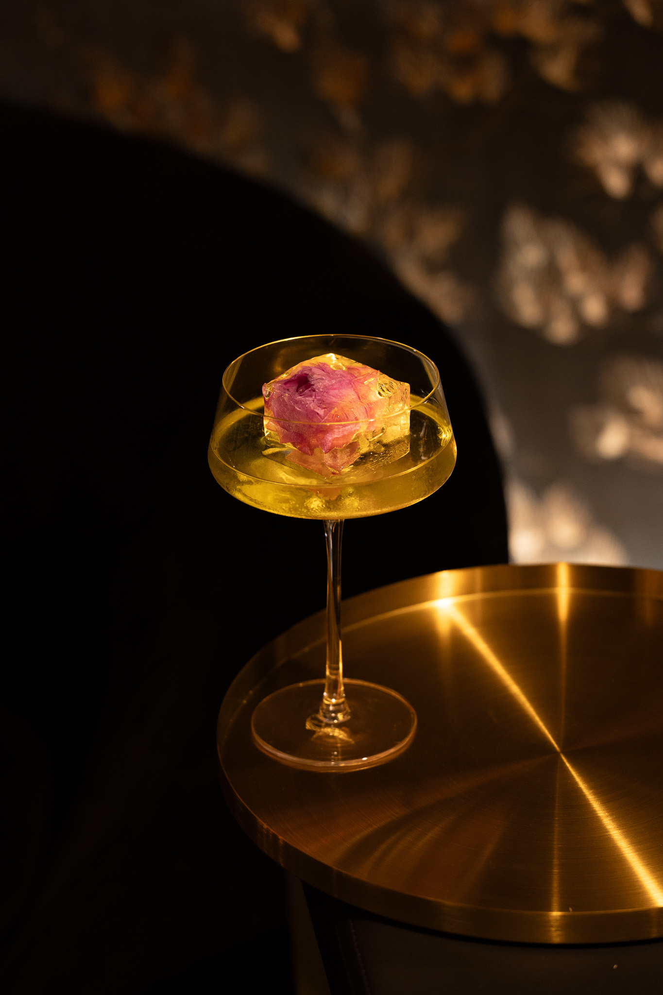 Elegant cocktail in a coupe glass with a large ice cube containing a pink flower, placed on a golden metallic table.