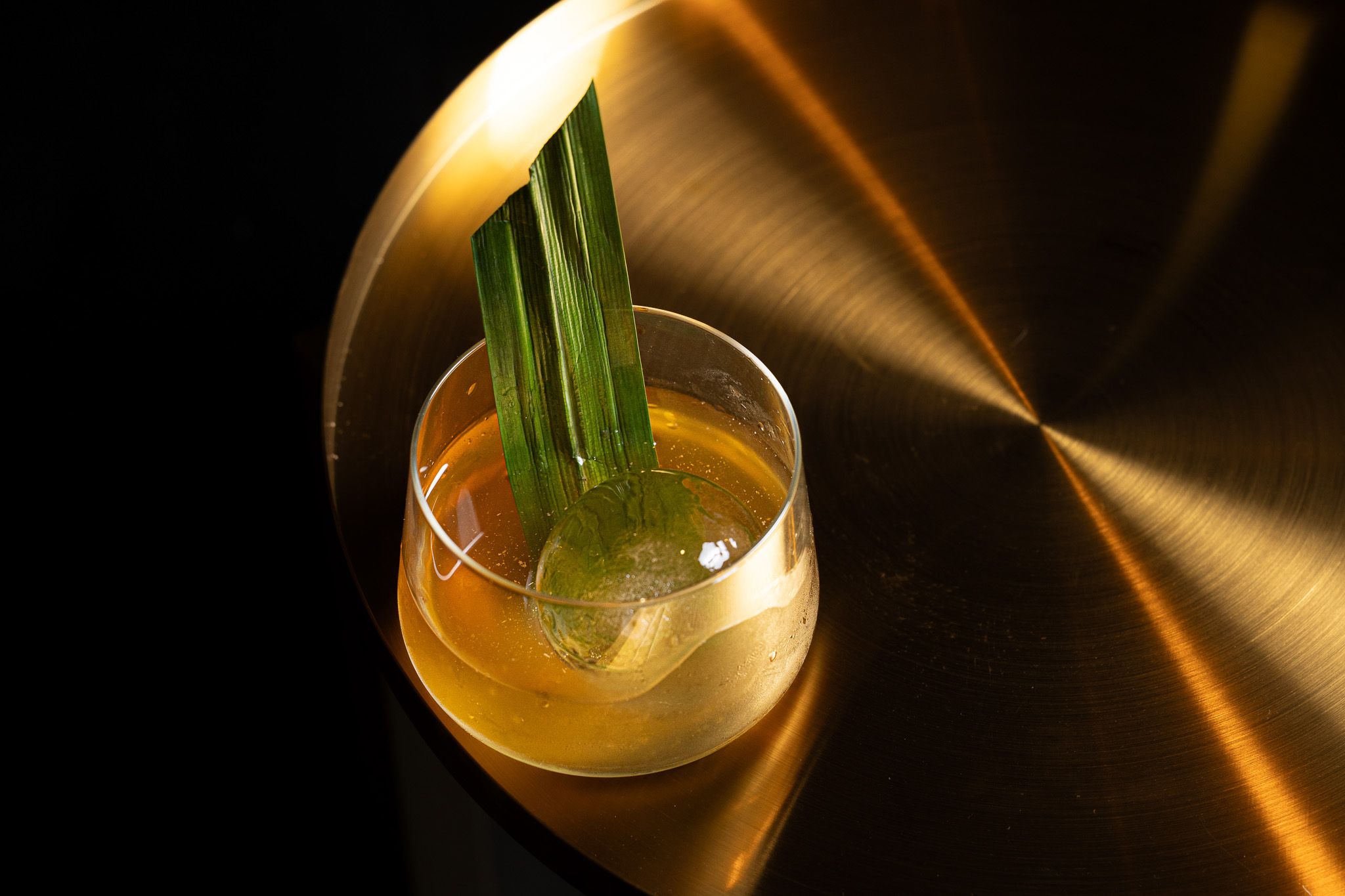 Cocktail with a large round ice sphere and two long green leaves in a short glass on a reflective bronze surface.