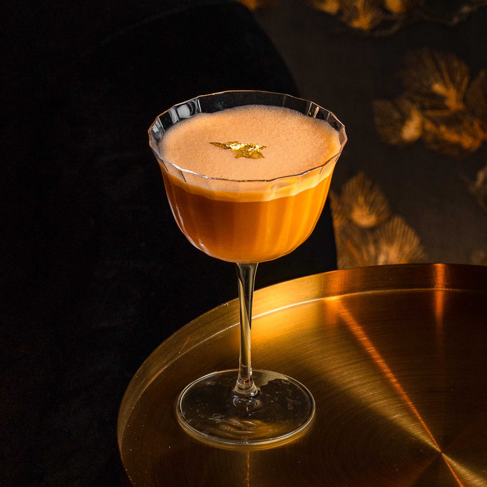 Foamy orange cocktail in a scalloped-edge coupe glass garnished with a small gold leaf, placed on a round metallic gold table.