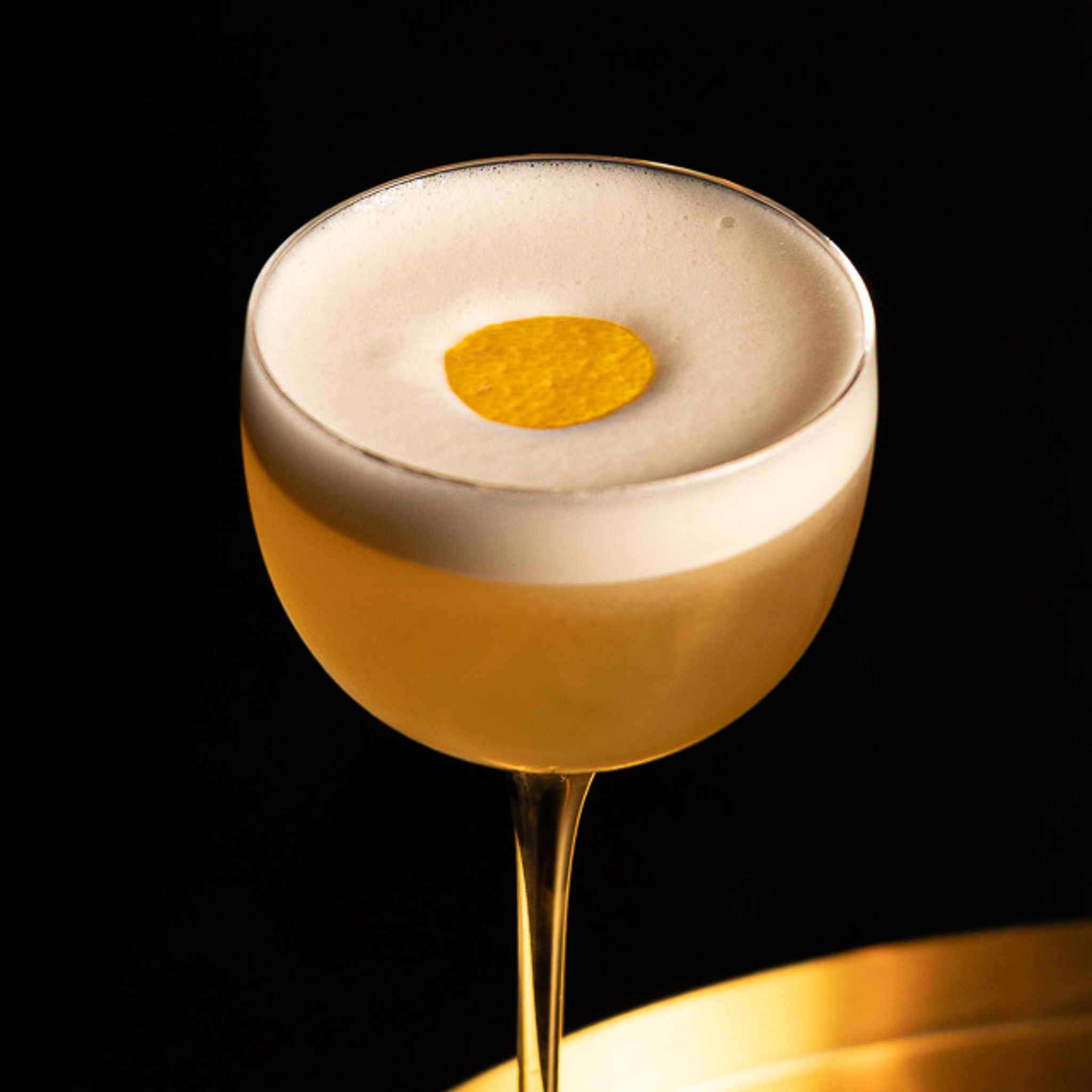 Cocktail in a coupe glass with frothy foam topped with a round orange garnish against a black background.