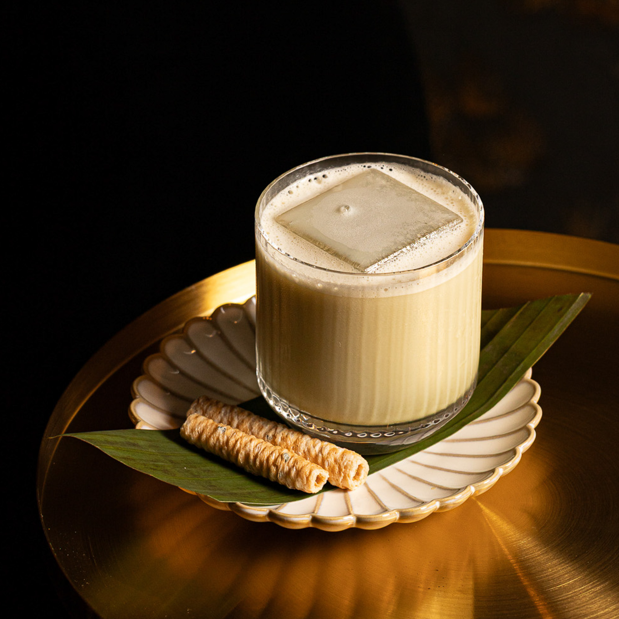 Creamy iced cocktail with a large square ice cube, served on a white scalloped plate with two wafer rolls on a green leaf.