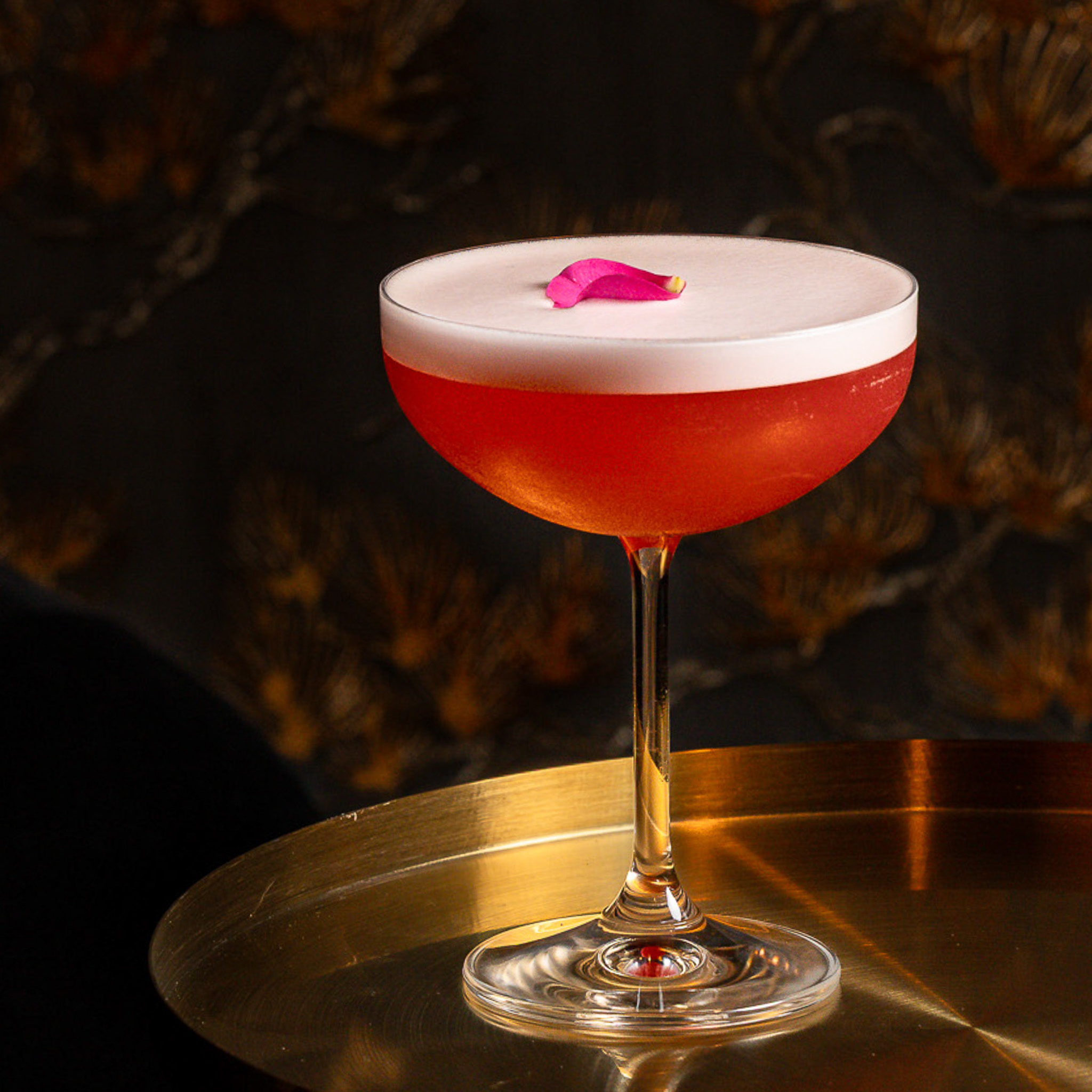 Elegant red cocktail with a frothy white top and pink flower petal garnish in a coupe glass on a reflective gold surface.