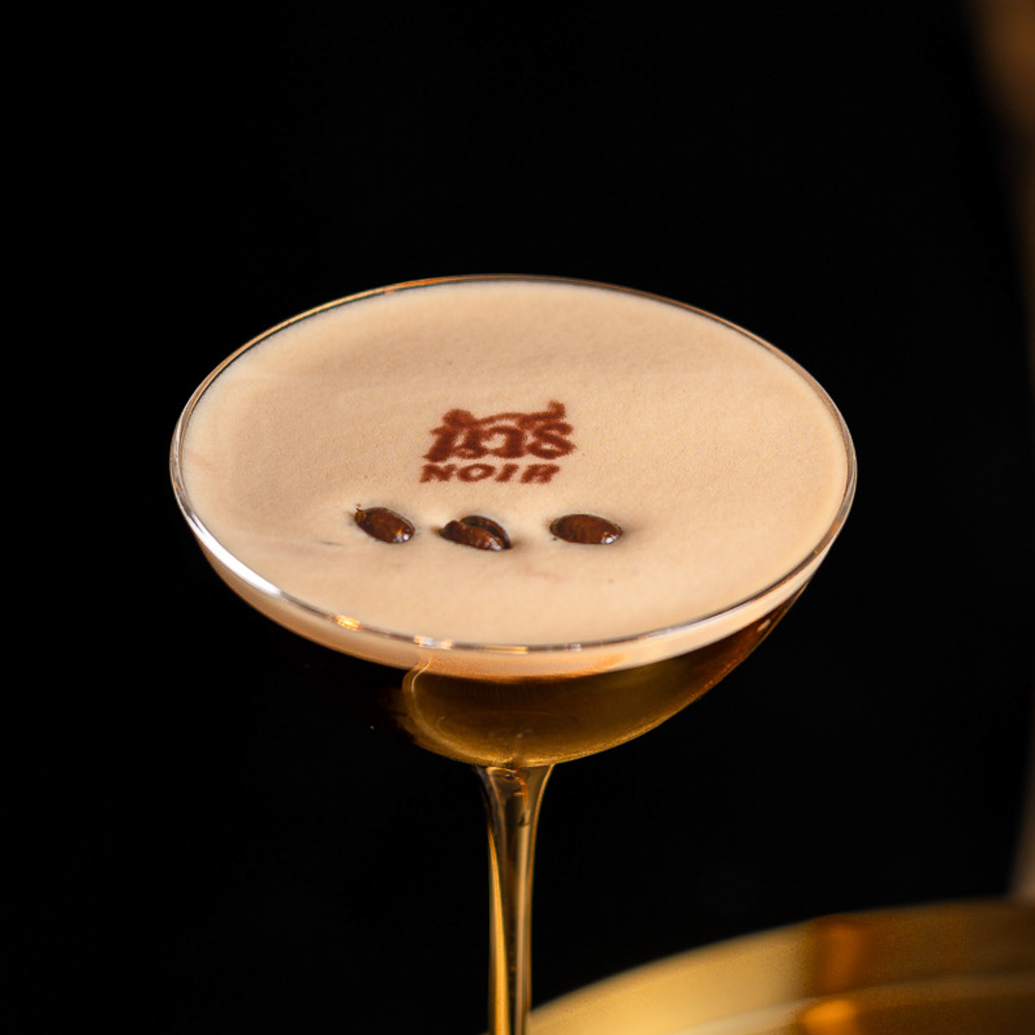 Cocktail glass with creamy coffee drink topped with three coffee beans and the word 'NOIR' printed in cocoa powder on the foam.