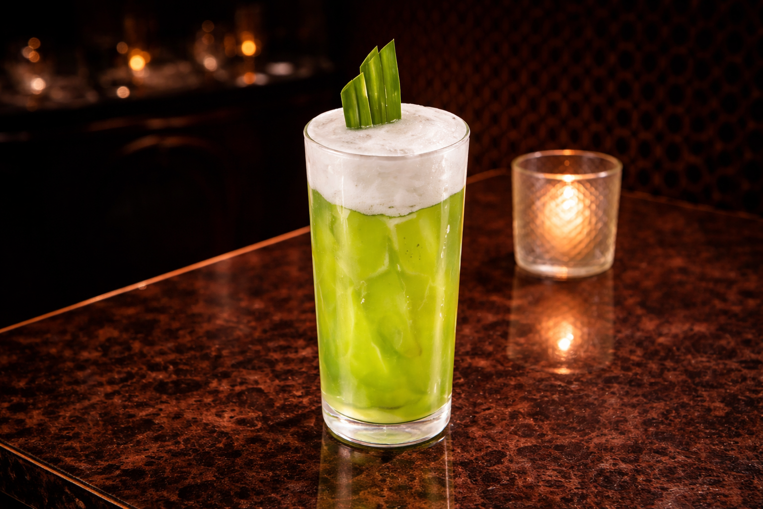Tall glass of iced green matcha latte topped with white foam and green leaf garnish on a brown marble table with candlelight in the background.