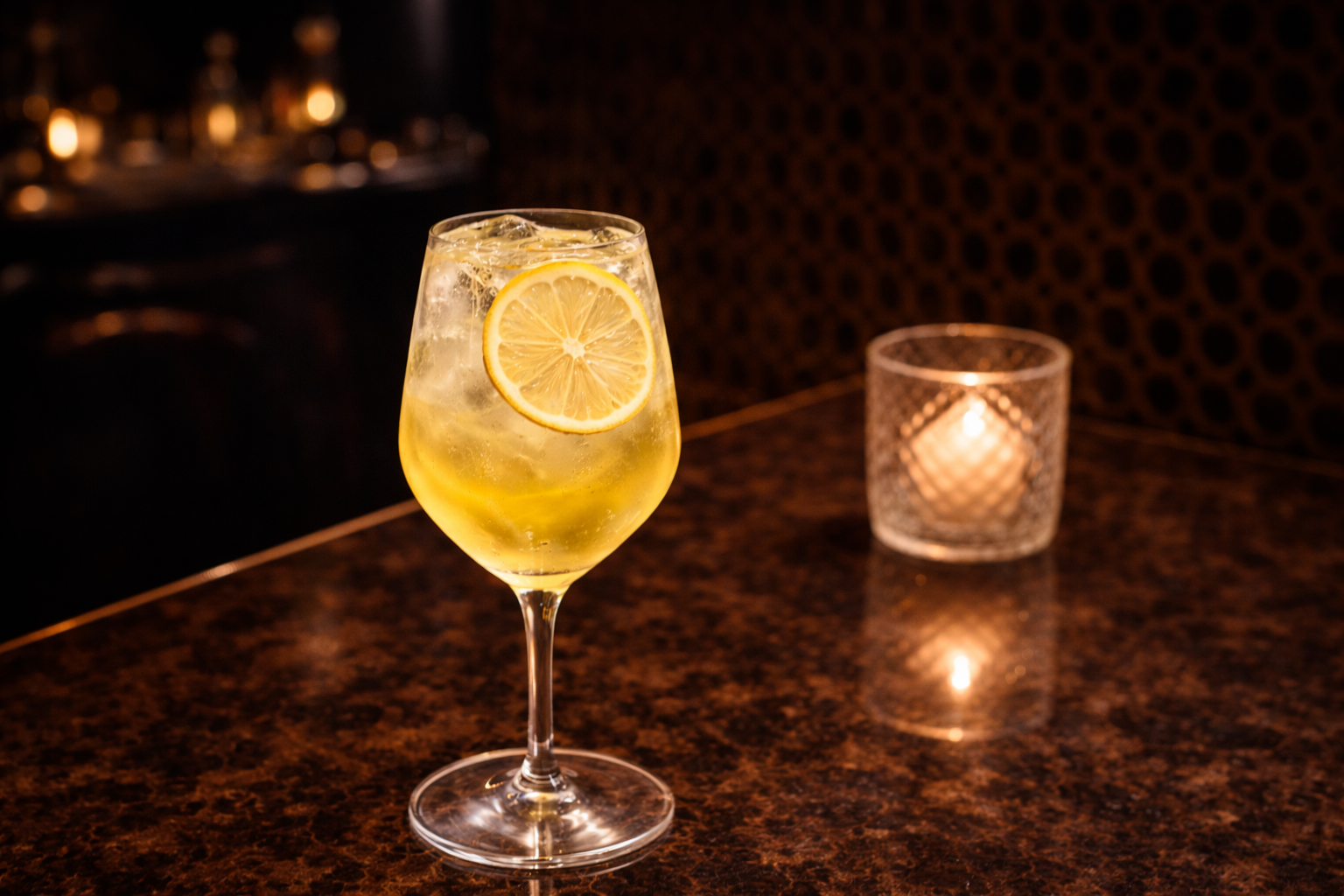 Glass of iced yellow cocktail garnished with lemon slice on a polished brown table with a lit candle in the background.
