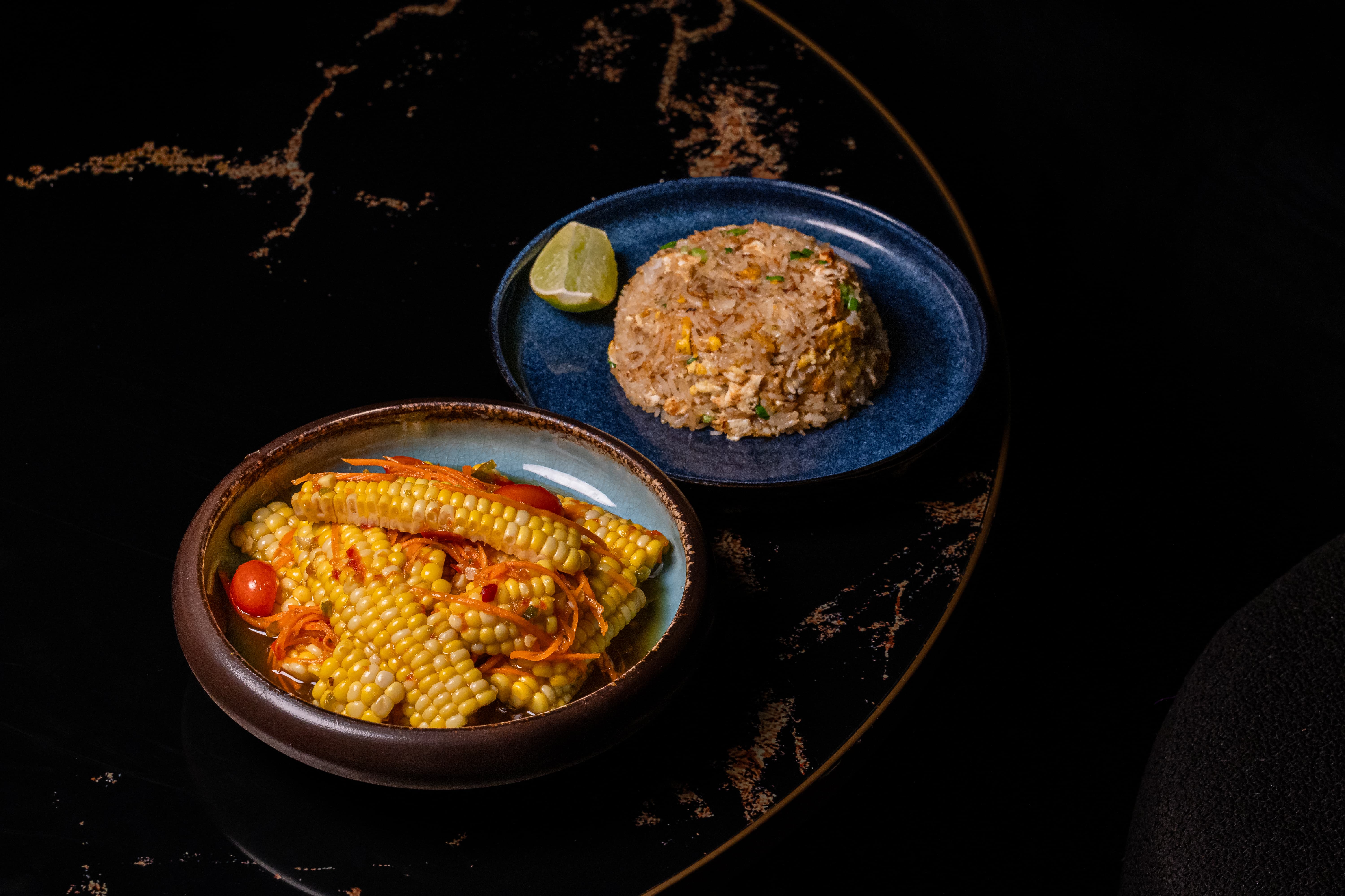 Bowl of corn with shredded carrots and cherry tomatoes alongside a plate with a mound of fried rice and a lime wedge.