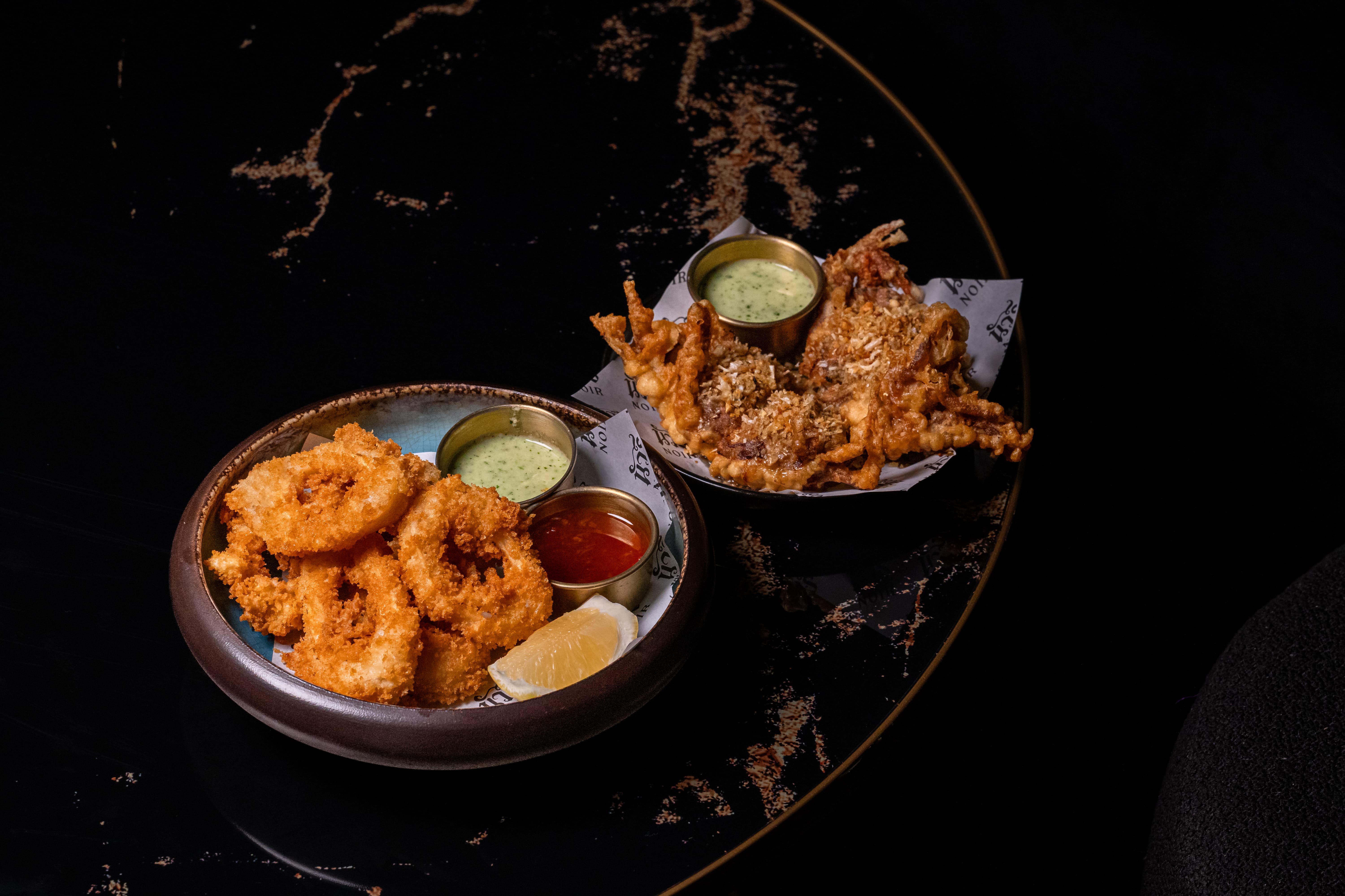 Two bowls with fried appetizers including golden-brown onion rings, fried soft-shell crab, lemon wedge, and dipping sauces on a dark table.