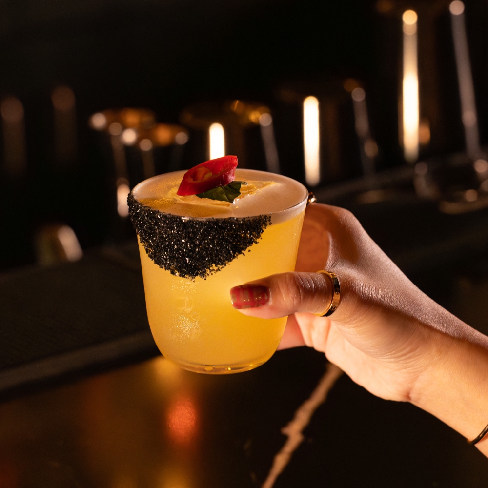Hand holding a yellow cocktail glass with black sugar rim and garnished with a red chili slice and green leaf.
