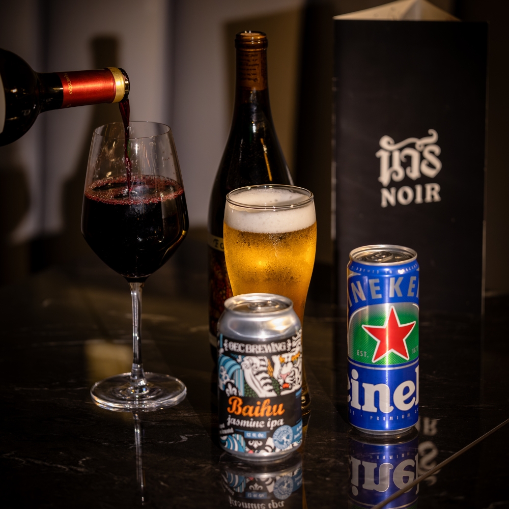 Red wine being poured into a wine glass, next to a glass of beer with foam on top, two beer cans, and a black menu labeled Noir.
