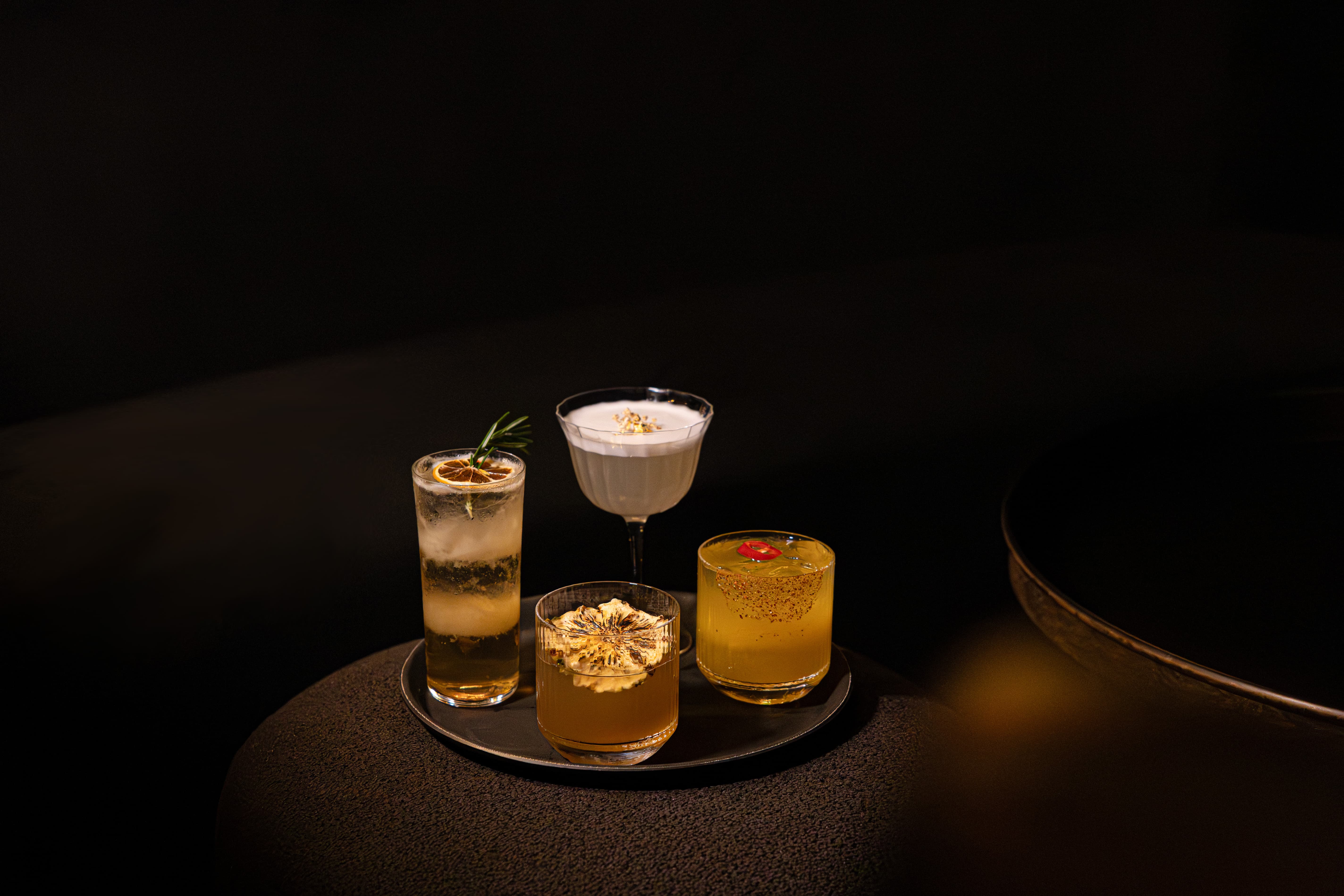 Four assorted cocktails on a black tray with dark background, each garnished uniquely with herbs, dried fruit, foam, or chili.