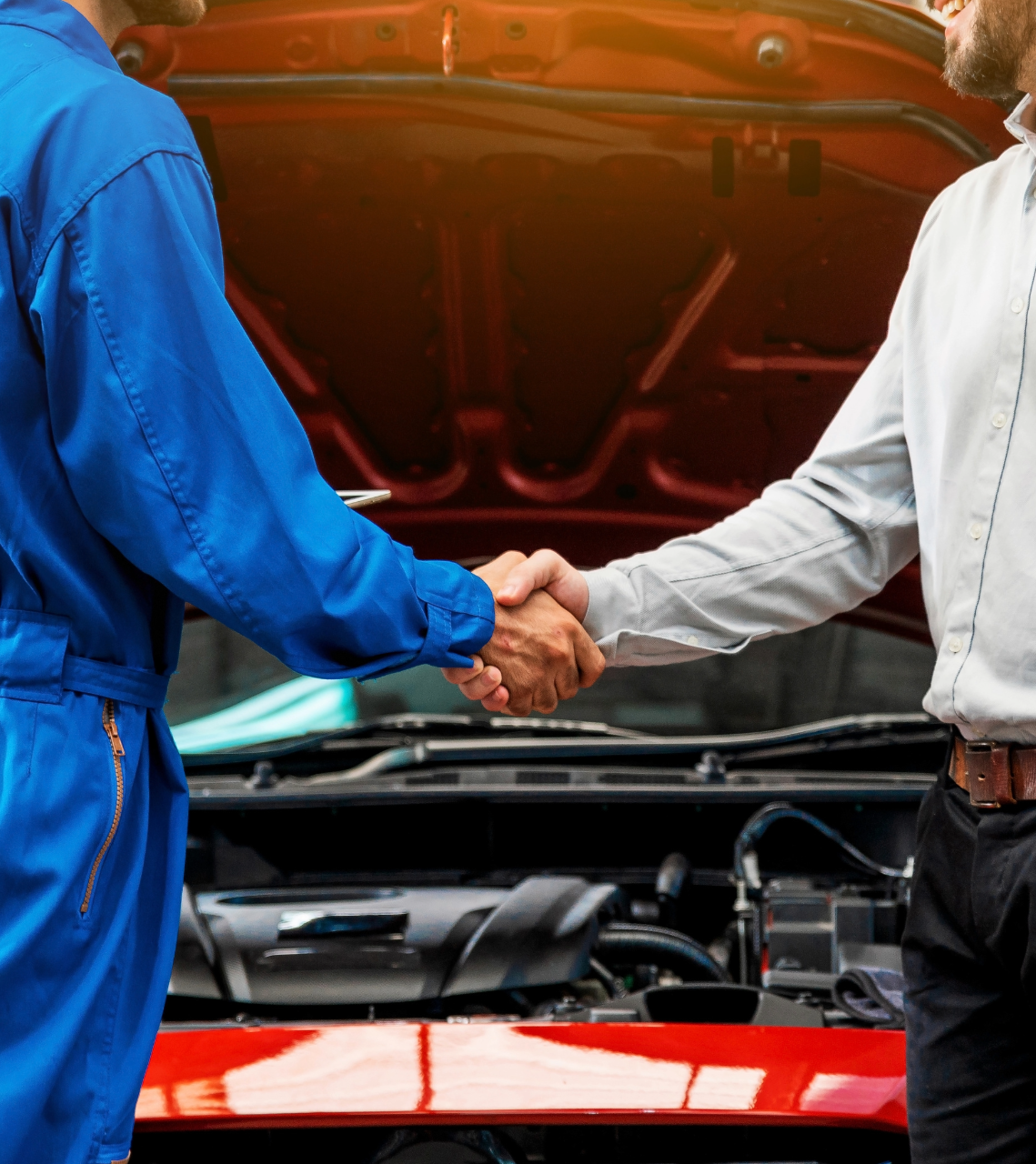 Mechanic in blue coveralls shaking hands with a smiling man in a white shirt in front of an open car hood.