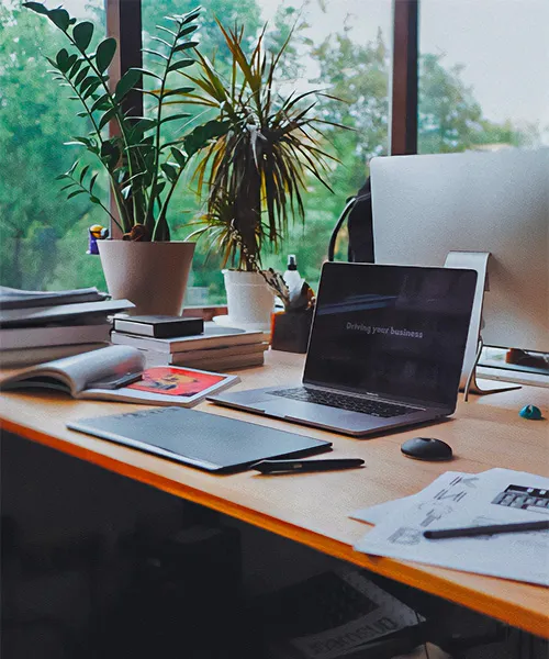 A laptop sits on a desk beside potted plants. The desk is beside a window with a view of trees.biophilic design