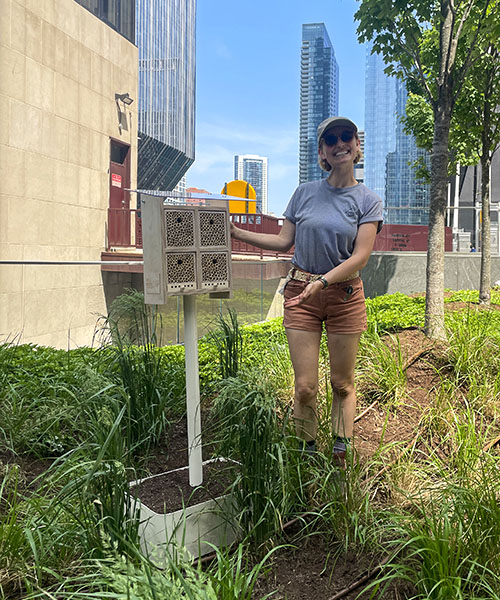 An Alvéole beekeeper smiles and gestures towards a BeeHome. They are standing on a green roof in the city, surrounded by modern sky scrapers.urban beekeeping benefits