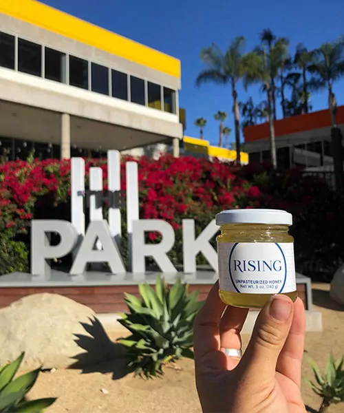 A hand holding a jar of honey with a custom label in front of the building where the honey was made. beekeping for commercial
