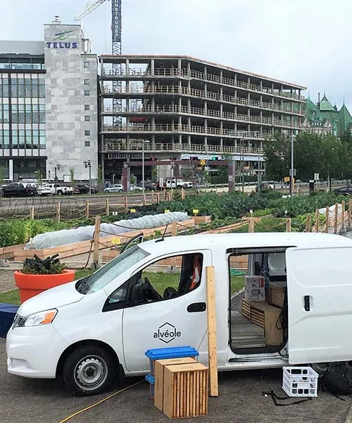 Une camionnette Alvéole remplie de matériel apicole stationnée devant un grand jardin urbain. agriculture urbaine