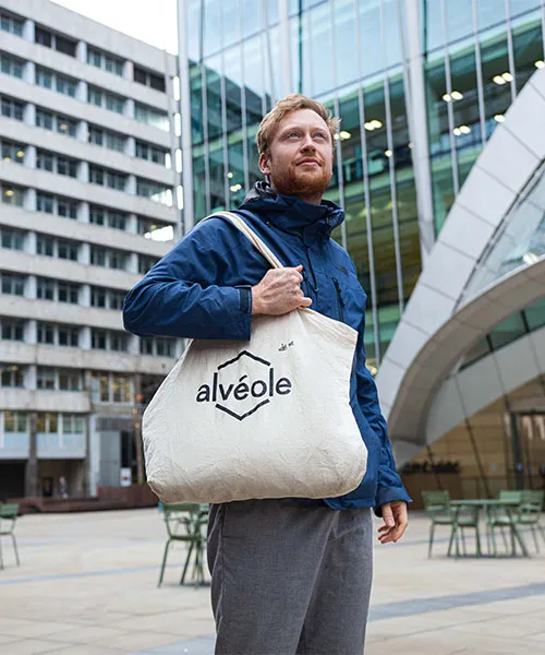 Un homme debout sur une place entourée de grands immeubles de verre, tenant un sac fourre-tout Alvéole. Alvéole B Corp