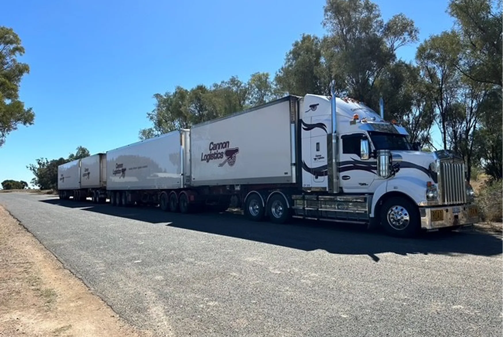 Large white Cannon Logistics semi-truck with three connected trailers parked on a road beside trees.