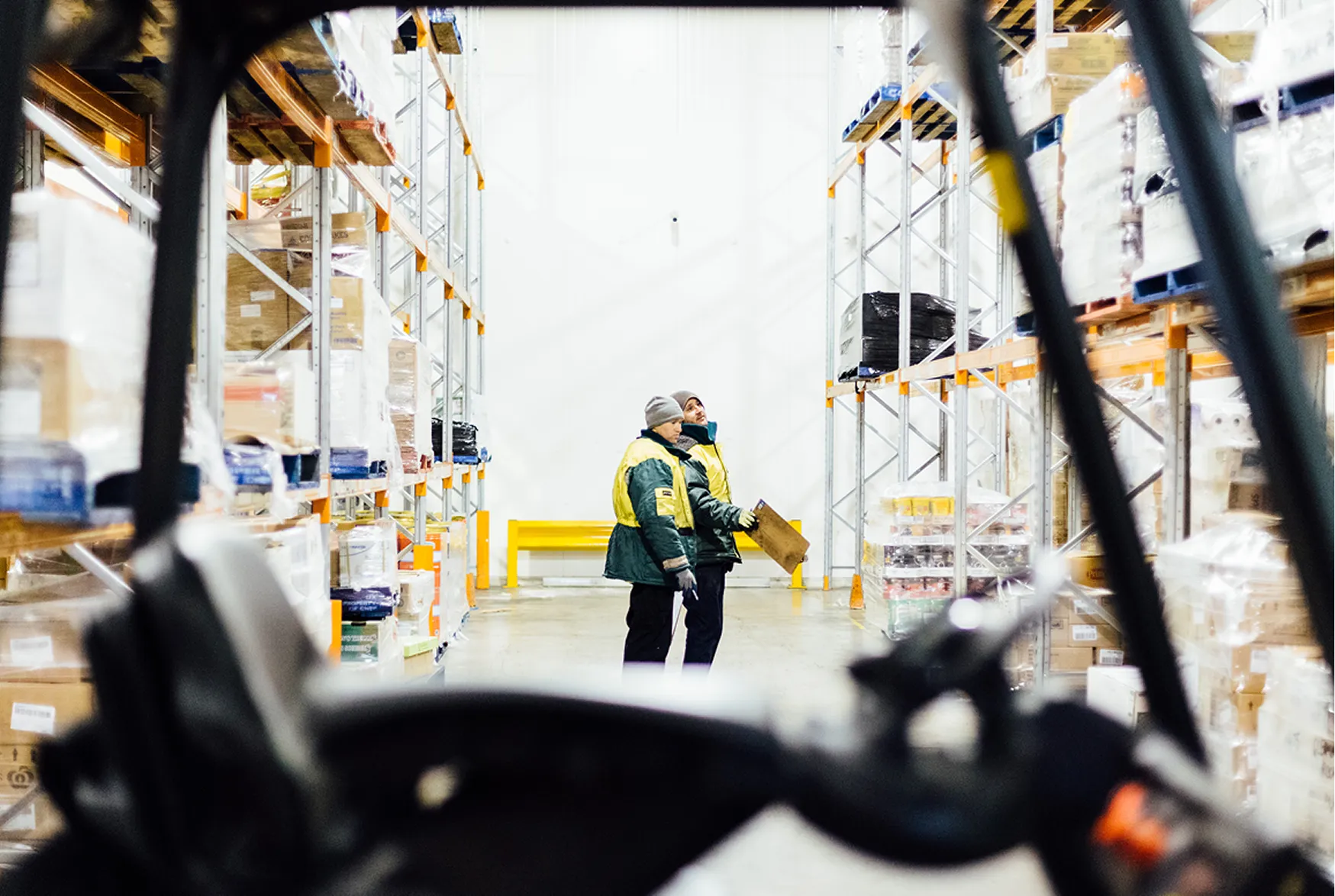 Two warehouse workers in yellow jackets inspecting storage shelves filled with boxes and pallets.