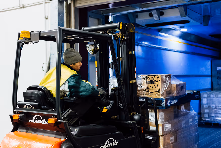 Worker in a yellow jacket and gray beanie operating a Linde forklift to load pallets in a warehouse.