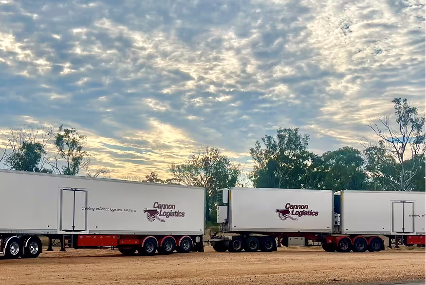 Two white trailers with Cannon Logistics logo parked on dirt with trees and cloudy sky in background.