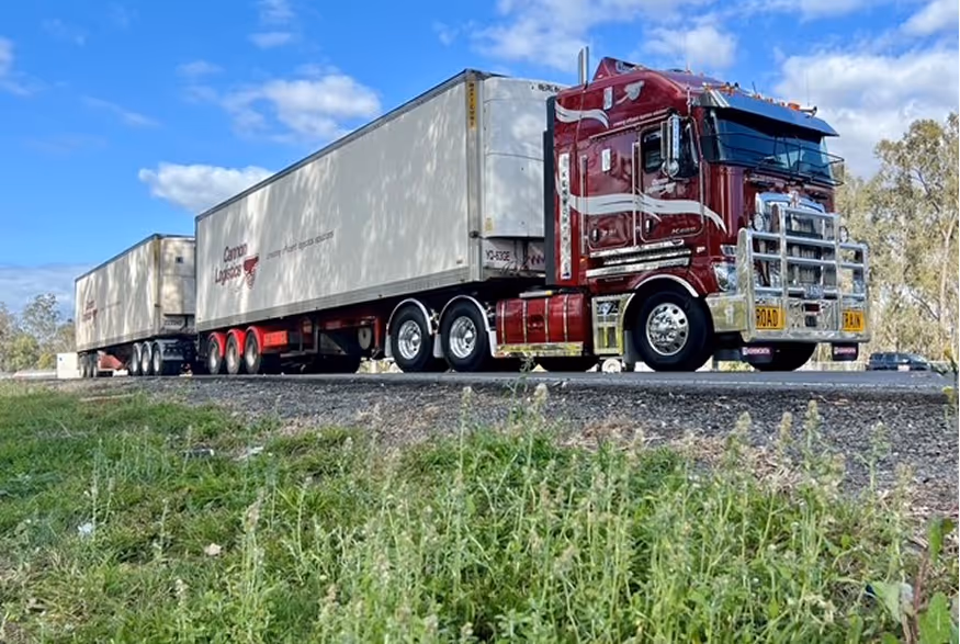 Red cab semi-truck with white double trailers on a road under a blue sky with clouds.