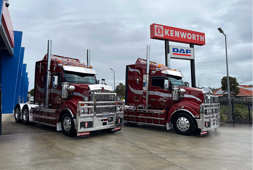 Two maroon Kenworth semi trucks parked outside a dealership with Kenworth and DAF signage.