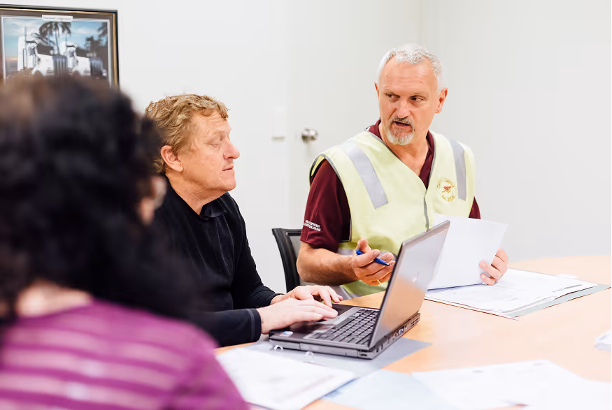 Two men in a meeting room discussing documents with a laptop on the table.