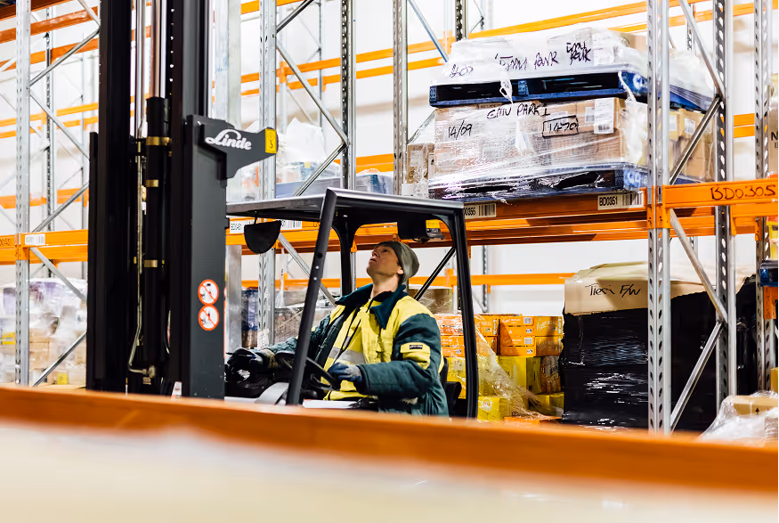 Warehouse worker operating forklift to lift pallet in racking system with boxes and packages.