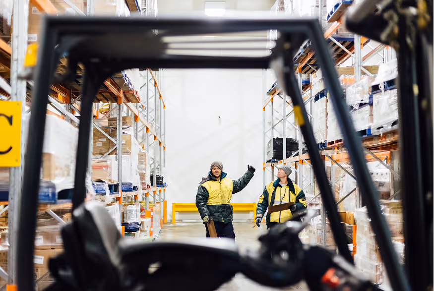 Two warehouse workers in yellow jackets talking amid storage shelves, viewed through forklift frame.