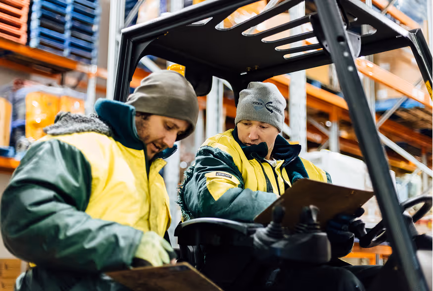 Two warehouse workers in yellow and green jackets reviewing clipboards near a forklift in storage racks.