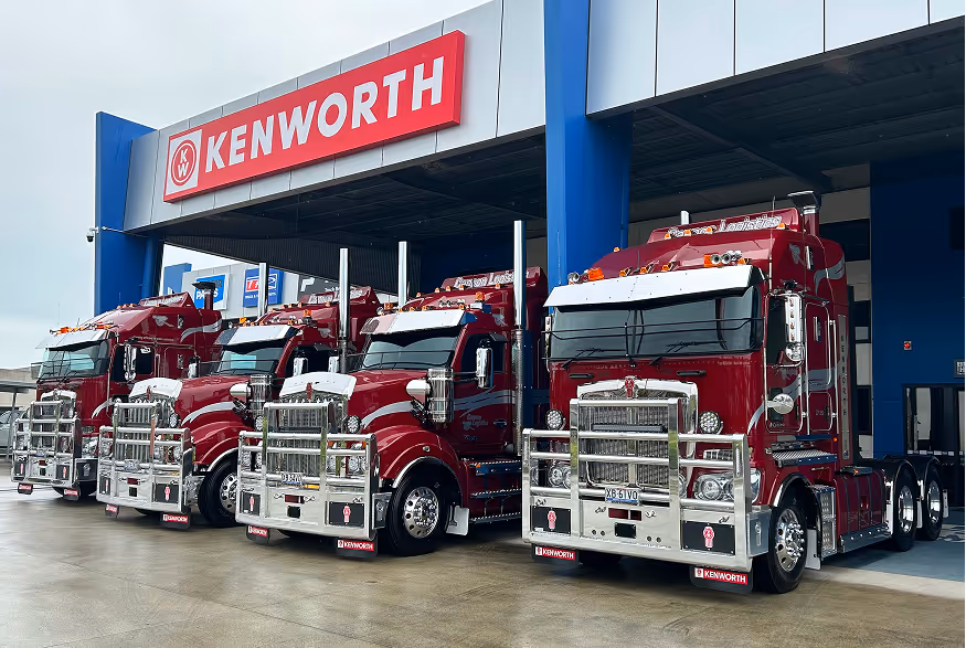 Four red Kenworth semi trucks parked in a row outside a Kenworth dealership.